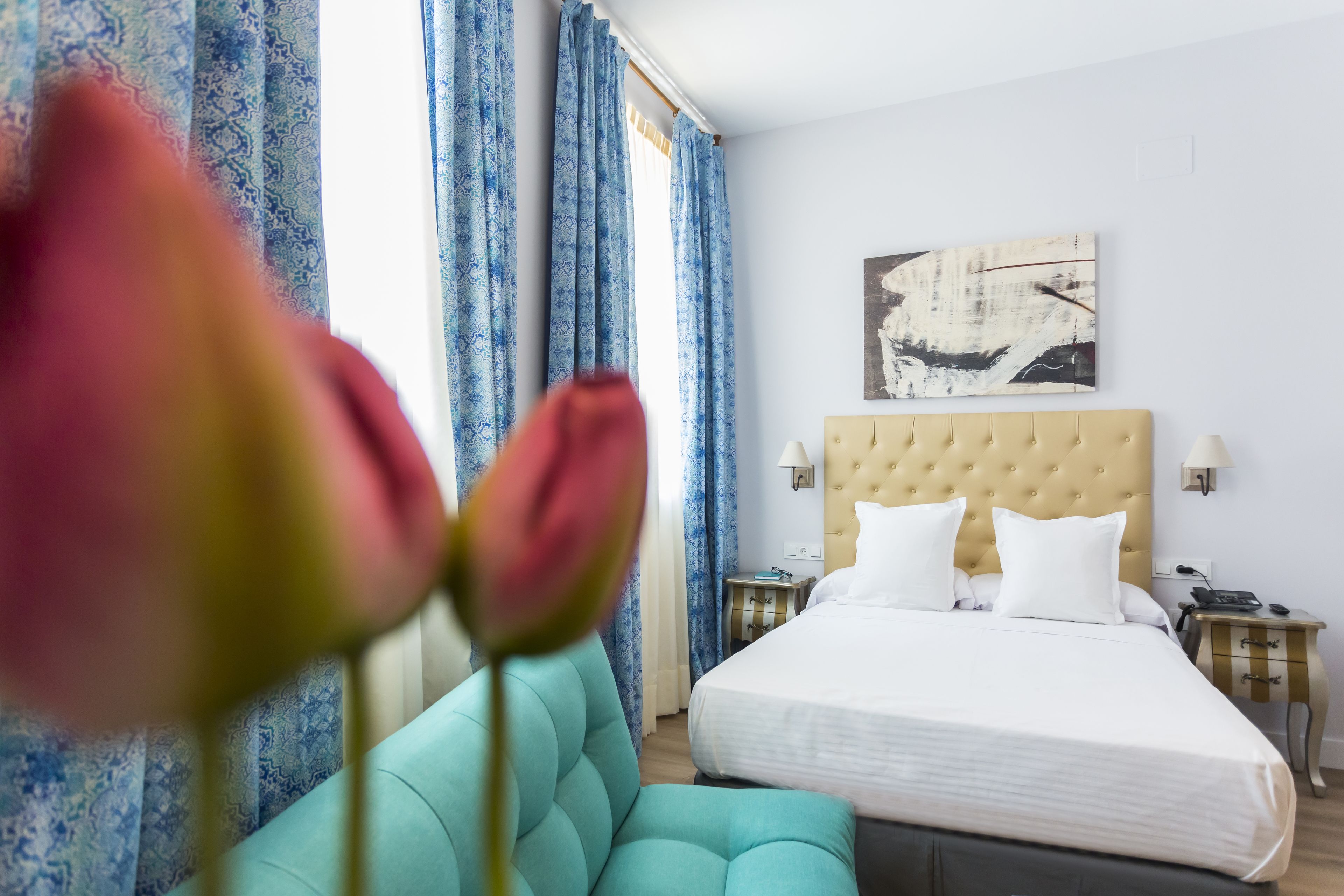 A hotel room with a large bed, a tufted headboard, a bedside table with a lamp, and a window with blue curtains.