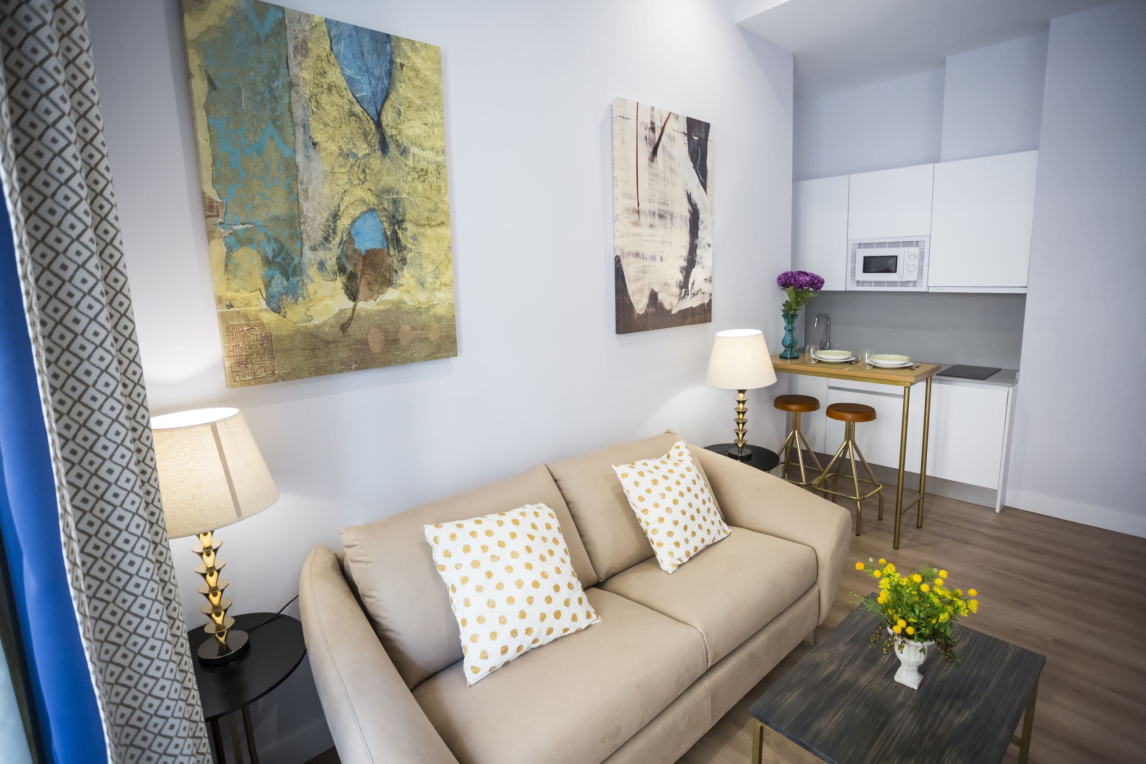 A modern living room with a beige sofa, patterned cushions, a small coffee table with a vase of yellow flowers, and a kitchen area with white cabinets and a microwave.