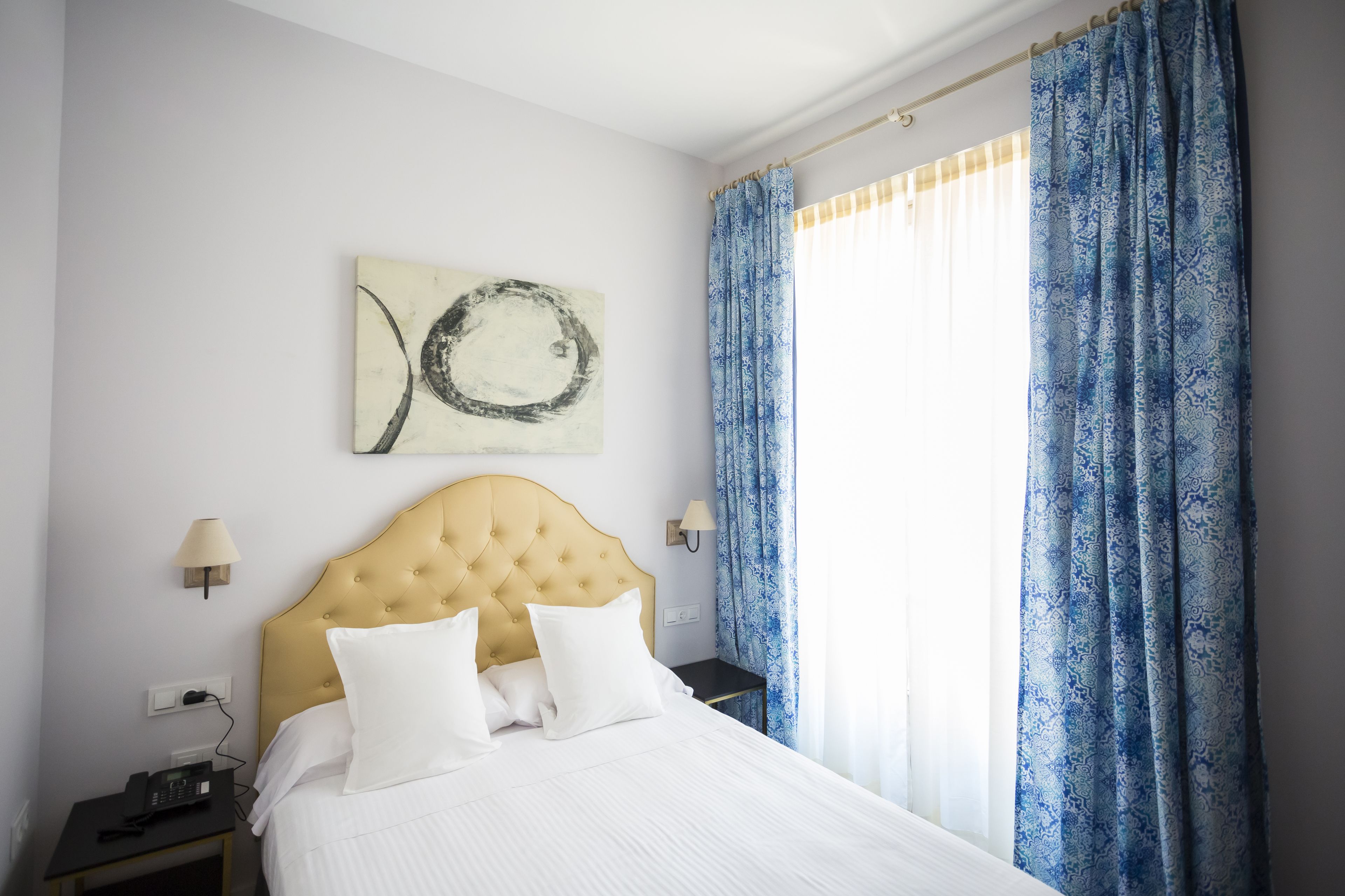 A hotel room with a bed, a nightstand, a lamp, a wall art piece, and a window with curtains.
