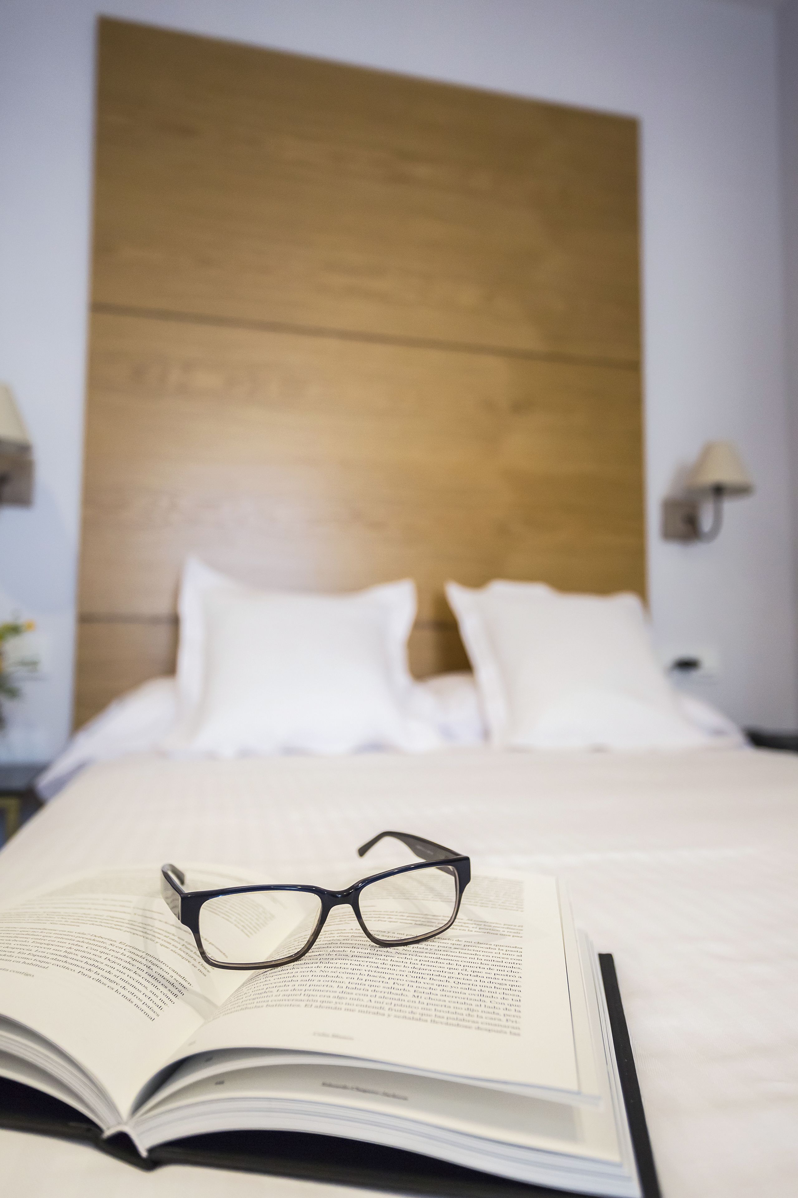 Una cama bien hecha con sábanas blancas, un cabecero de madera y un par de gafas sobre un libro abierto.