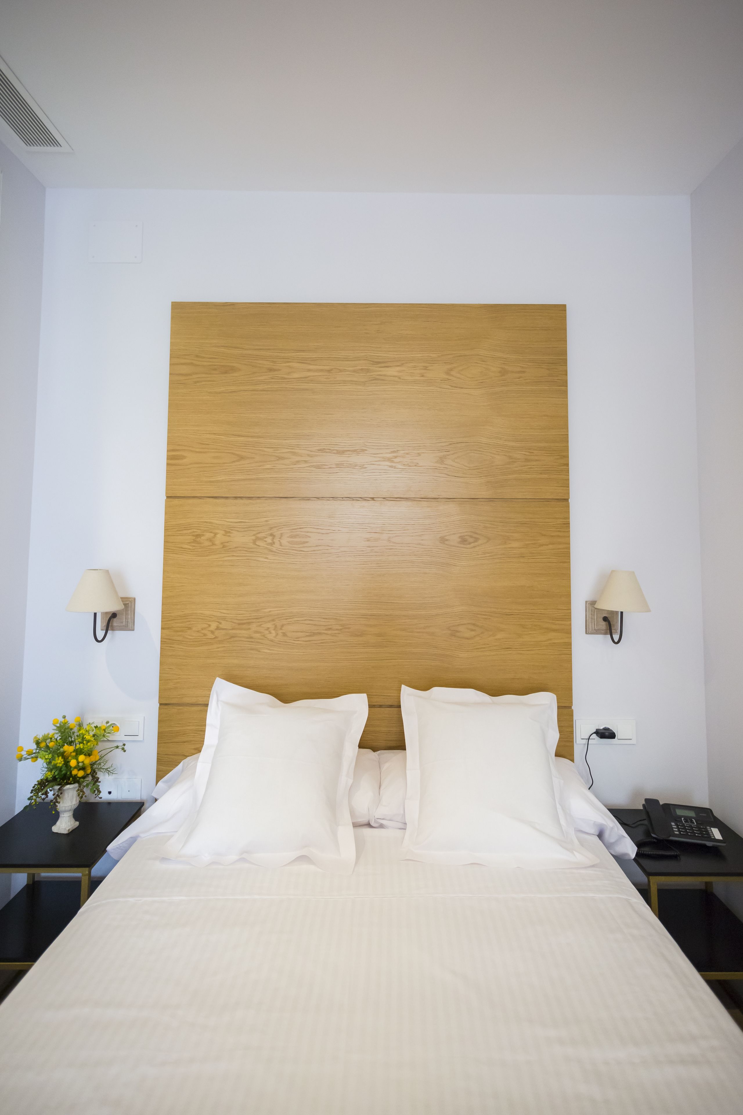 A hotel room with a wooden headboard, two bedside tables with lamps, a bed with white bedding, and a vase with yellow flowers on one table.