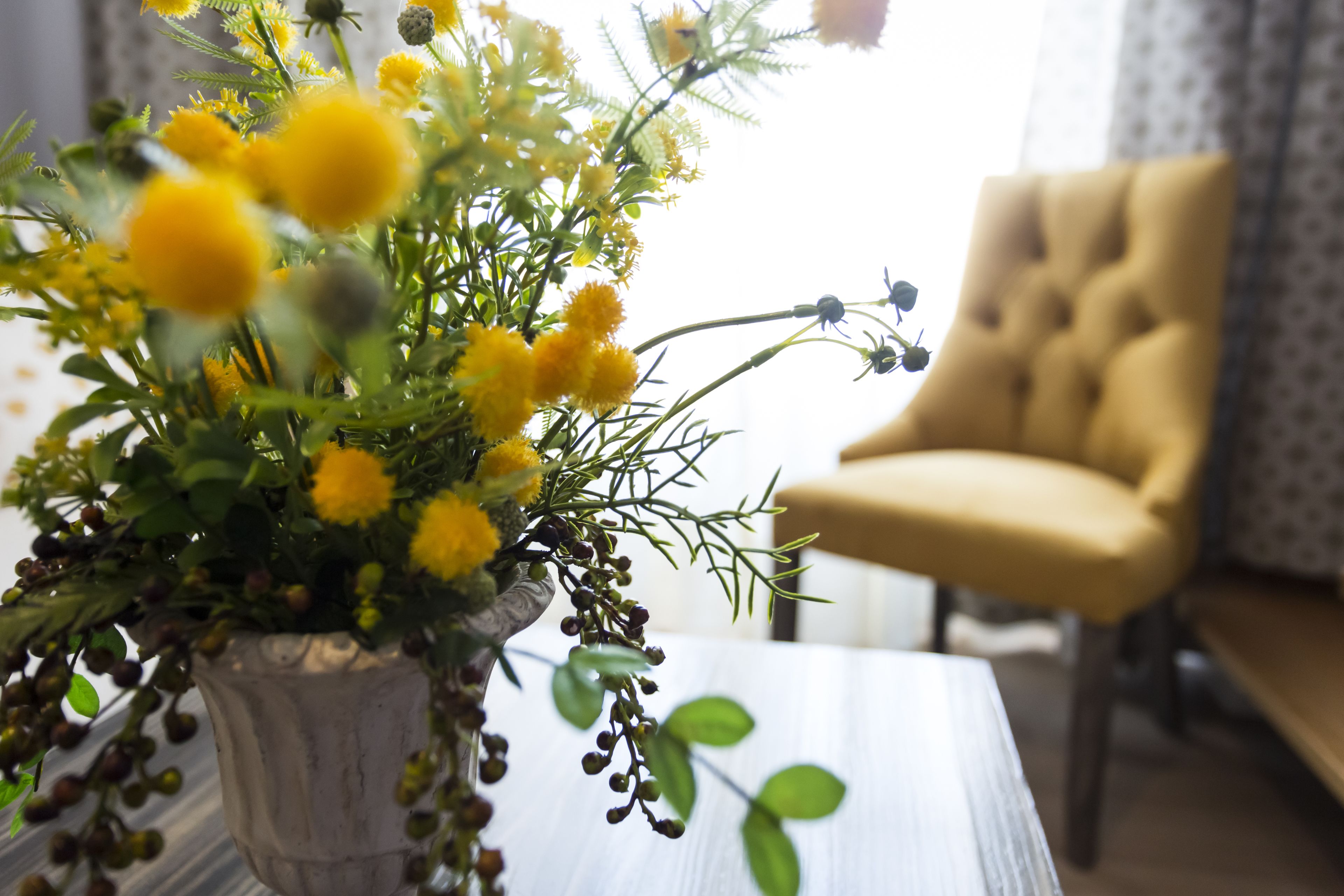 A room with a wooden table, a vase with yellow flowers, and a beige armchair.