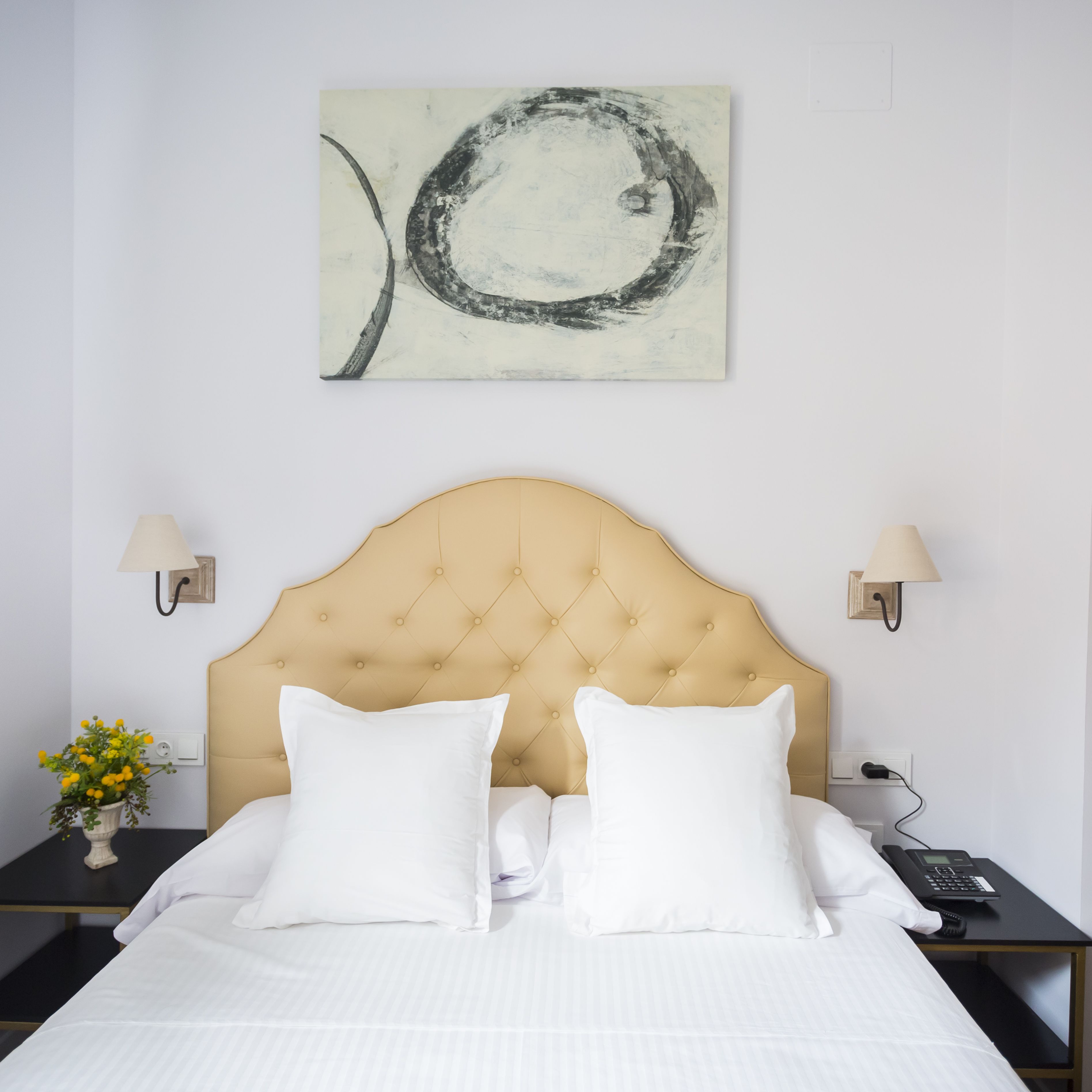 Ein Hotelzimmer mit einer gepolsterten Kopfteile, weißer Bettwäsche, einem einzelnen Kunstwerk an der Wand und einem Nachttisch mit Lampe und Telefon.