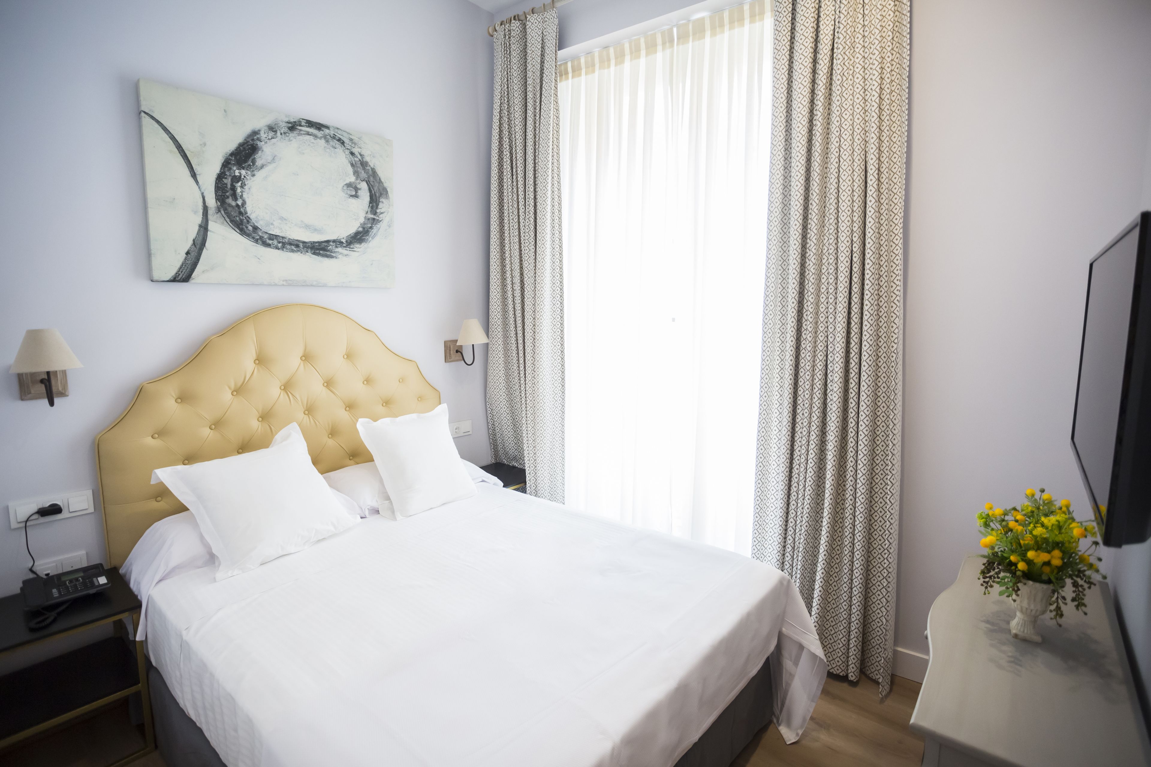 Habitación de hotel con una cama grande, cabecera, mesita de noche con lámpara, televisor montado en la pared y una ventana con cortinas.