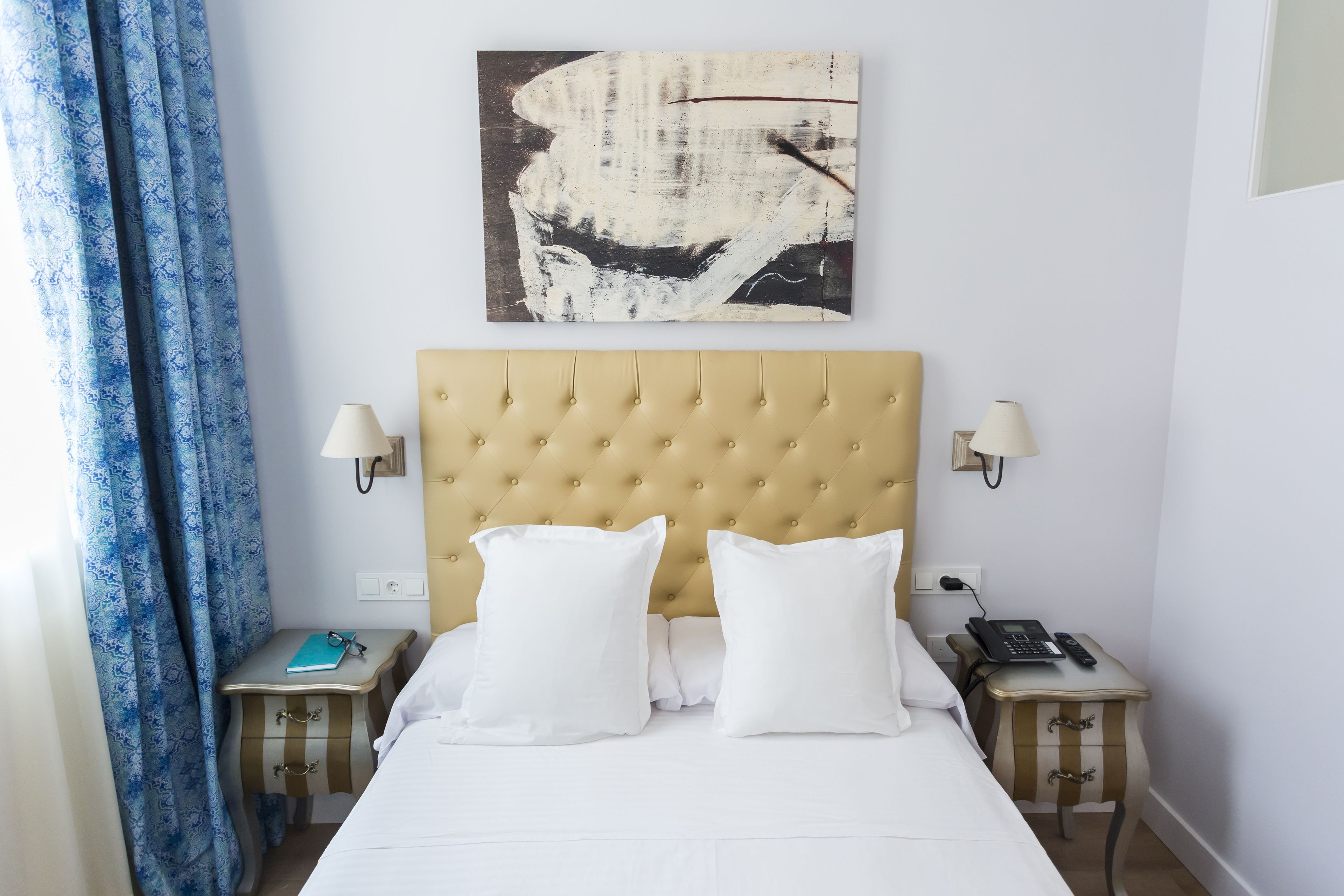 A hotel room with a tufted headboard, two bedside tables with lamps, a bed with white linens, a blue curtain, and a painting on the wall.