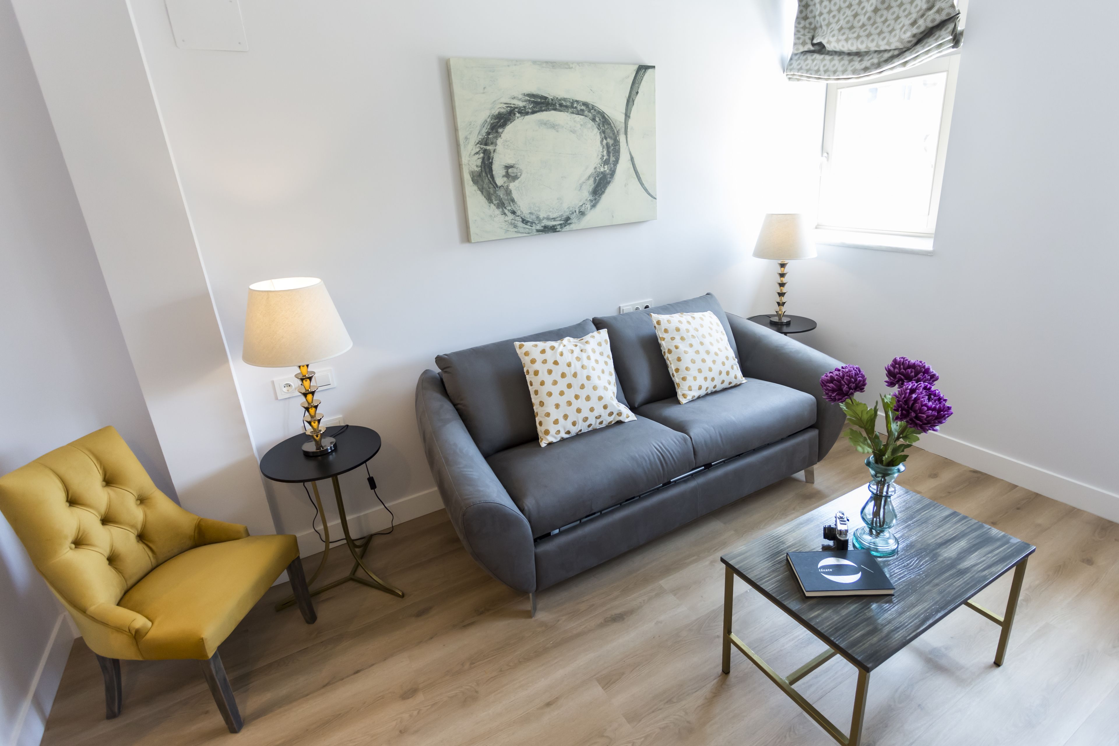 A modern living room with a grey sofa, a yellow armchair, a small round table, and a coffee table. There is a painting on the wall and a window with curtains.