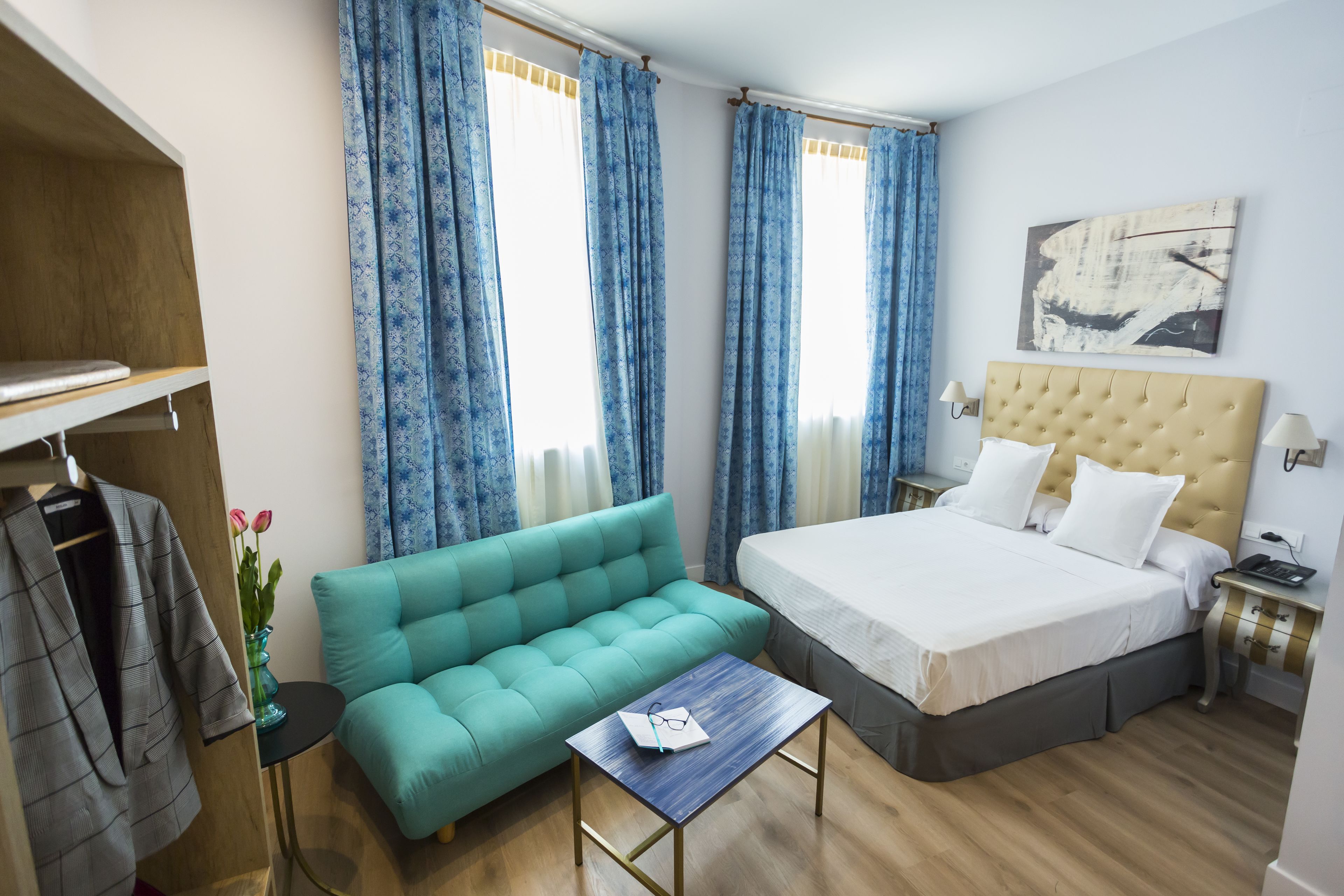 Una habitación de hotel con cama, sofá, una mesita, armario y ventana con cortinas.
