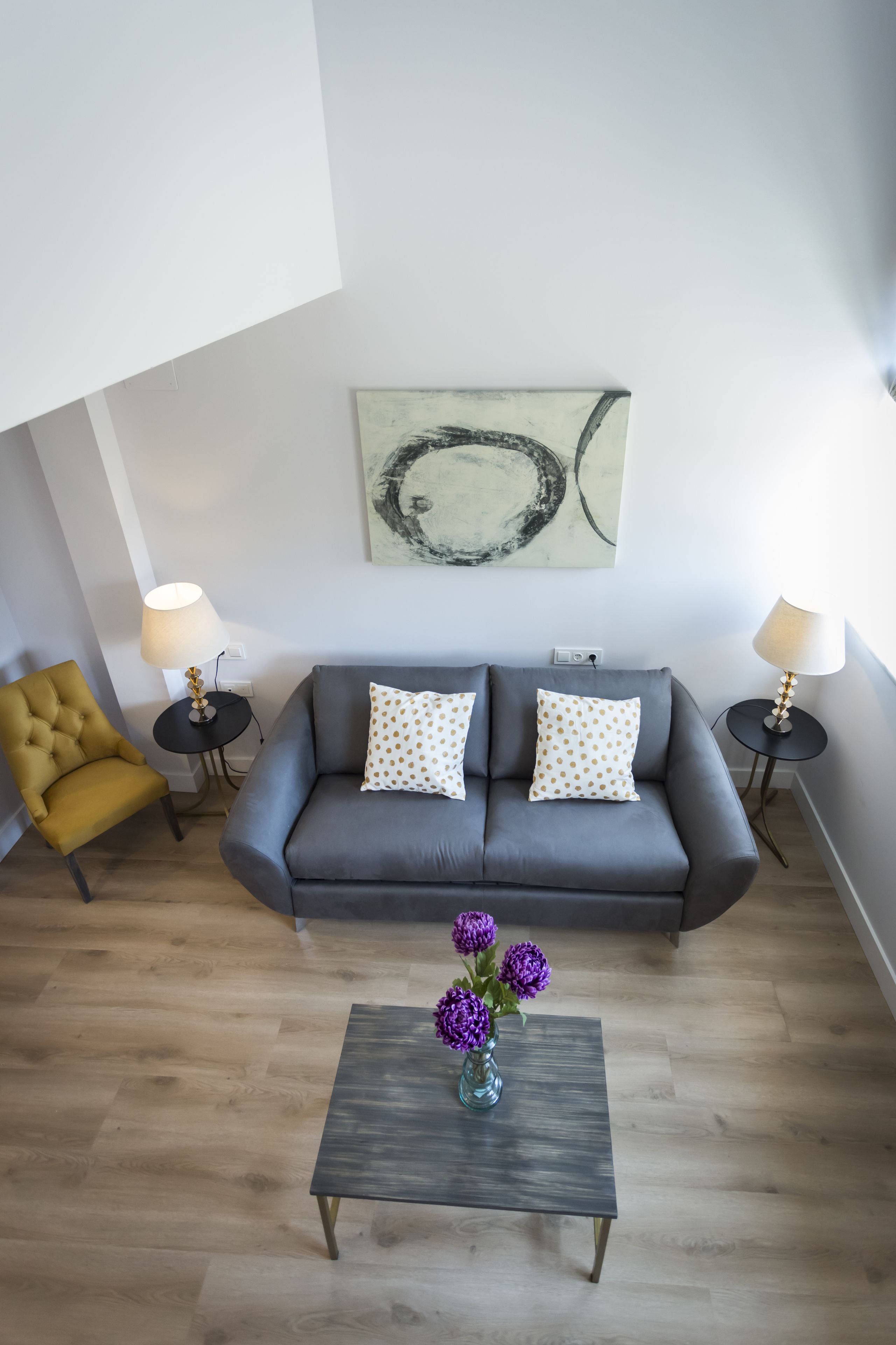 A modern living room with a grey sofa, two side tables with lamps, a mustard-colored armchair, and a coffee table with purple flowers.