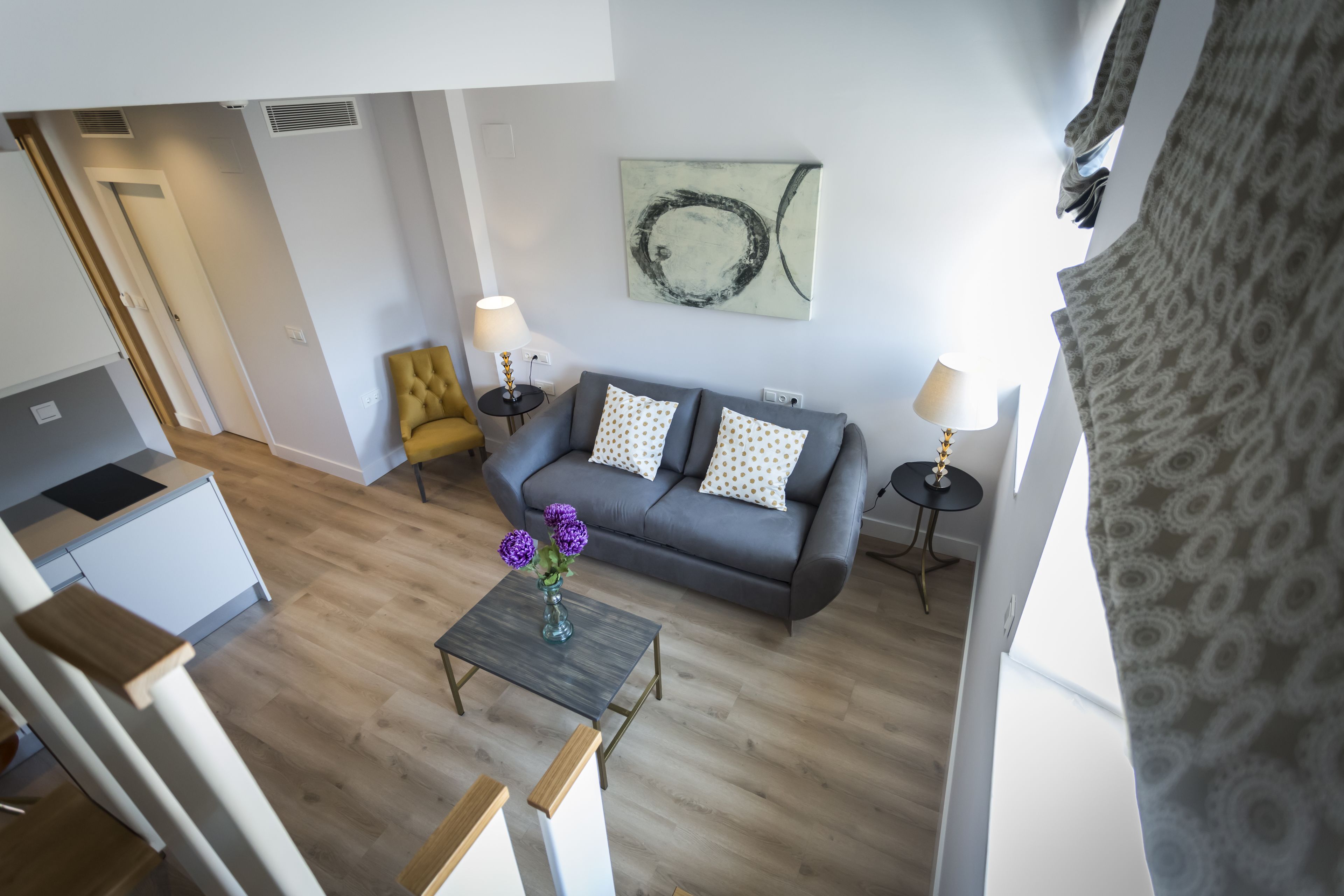 Een moderne woonkamer met een grijze bank, twee bijzettafels met lampen, een klein bijzettafeltje met bloemen en een keuken in de achtergrond.