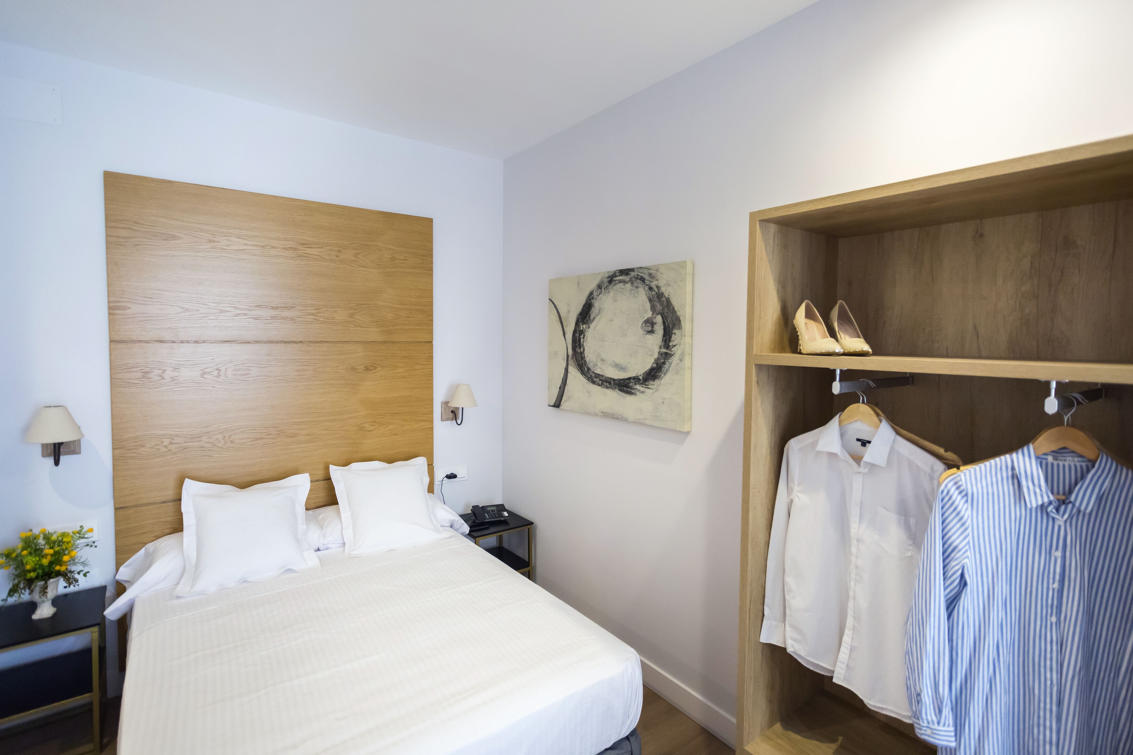 A hotel room with a large bed, a wooden headboard, a small bedside table with a lamp, a wall-mounted artwork, and a clothing rack with shoes and shirts.