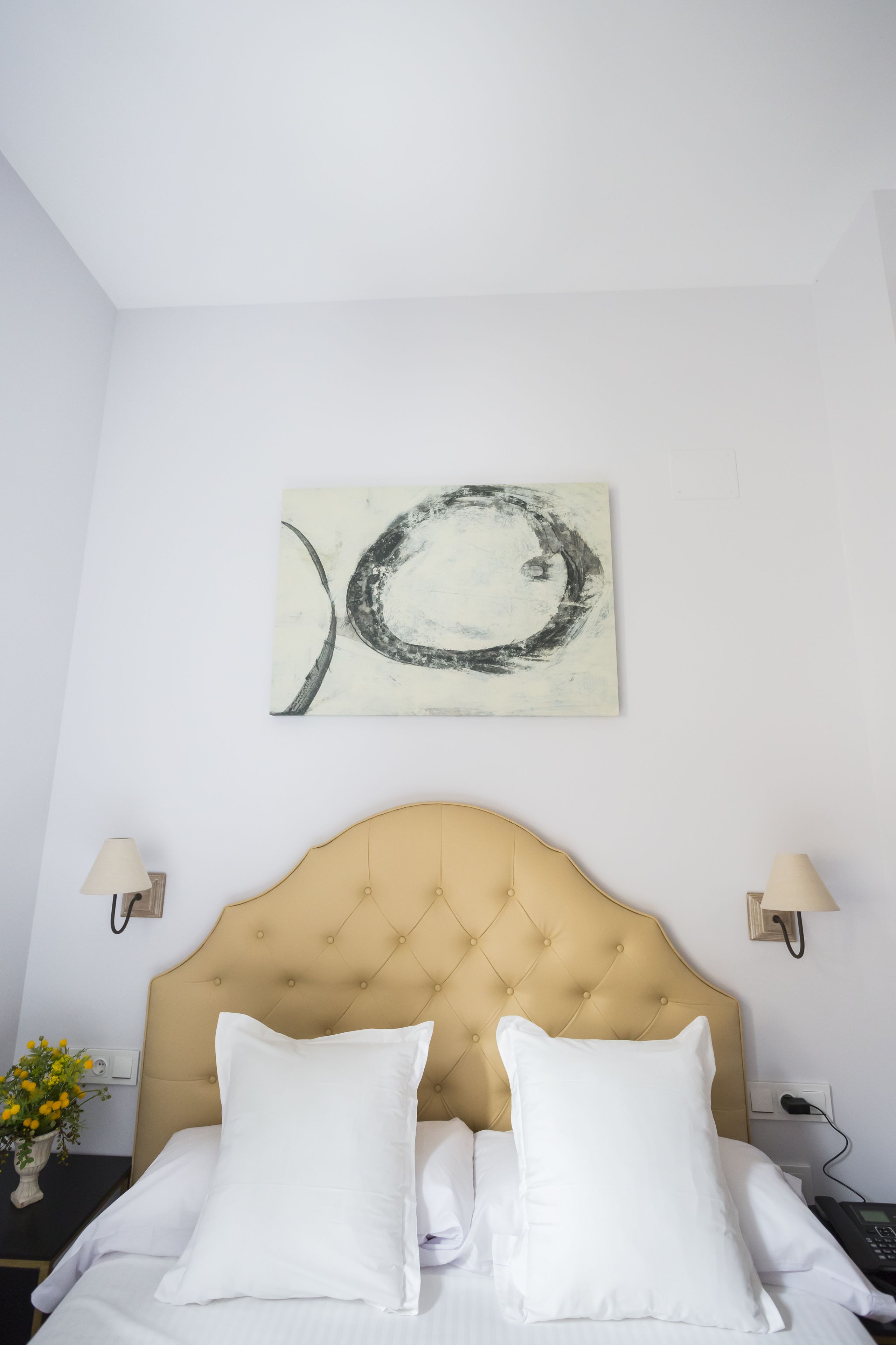 A hotel room with a tufted headboard, white bedding, a wall-mounted abstract artwork, bedside lamps, and a small table with a vase of flowers.