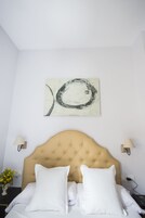 Habitación de hotel con cabecera capitonada, ropa de cama blanca, una obra de arte abstracta colgada en la pared, lámparas de noche y una mesita con un florero.