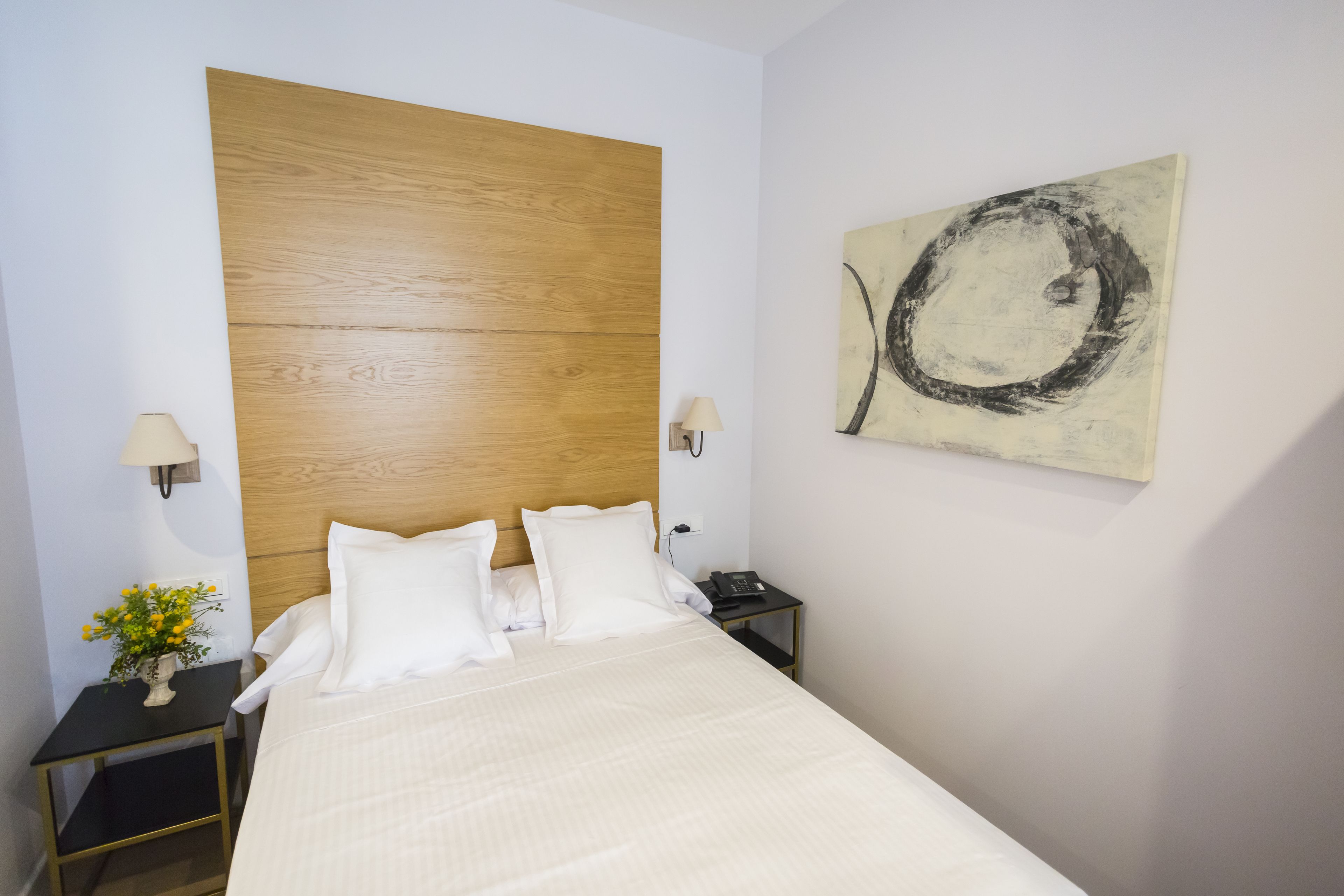 Une chambre d’hôtel avec une tête de lit en bois, un lit avec du linge de lit blanc, une table de chevet avec une lampe, un téléphone et une œuvre d’art murale.