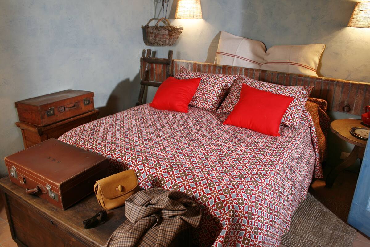 Ein Bett mit einem gemusterten Überwurf, zwei rote Kissen, einem Holz-Kopfteil und einem Nachttisch mit einer Lampe.