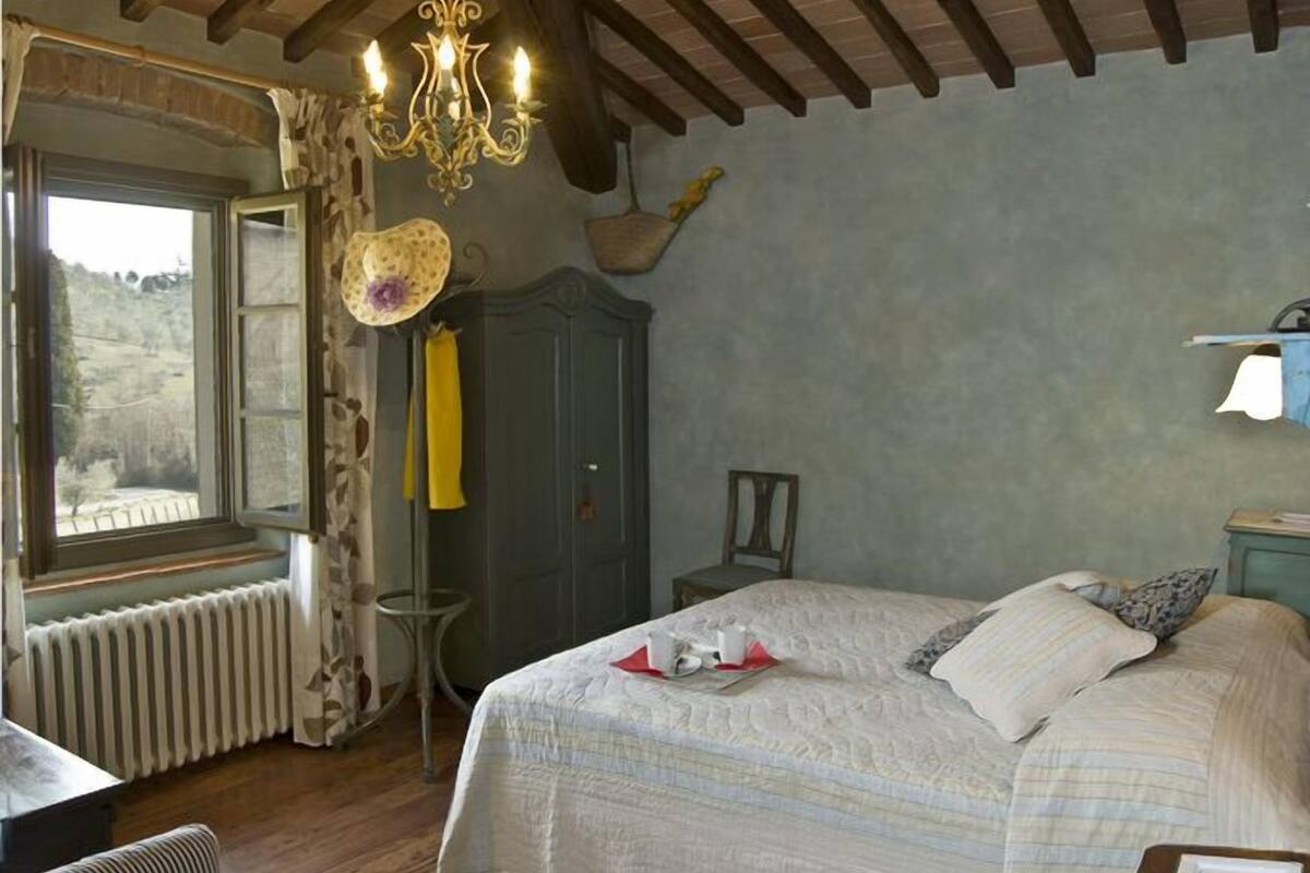 Ein Schlafzimmer mit einem großen Bett, einer Kristallleuchter, einem Fenster mit Vorhängen, einer Heizung, einem Holzboden und einem kleinen Tisch mit einer Blume.
