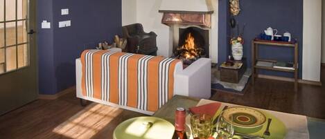 A cozy living room with a fireplace, a sofa, and a dining table set with a bottle, glasses, and fruit.