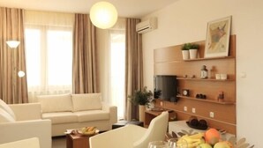 Una habitación de hotel moderna con sofá, una mesa de comedor con sillas, un televisor montado en la pared y un estante con objetos decorativos.