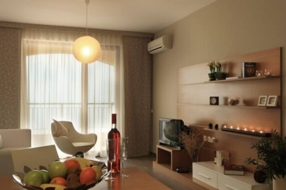 Ein modernes Wohnzimmer mit einer Couch, einem Esstisch mit Früchten und einer Weinflasche, einem Fernseher und einem Bücherregal.
