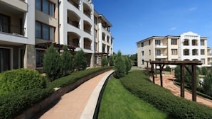 Un moderno edificio residencial de varios pisos, con balcones, un jardín ajardinado y un paseo pavimentado.