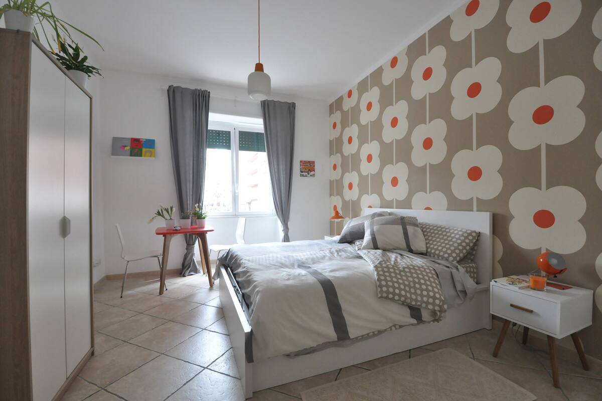 A bedroom with a bed, a bedside table, a chair, a desk, and a window with curtains.