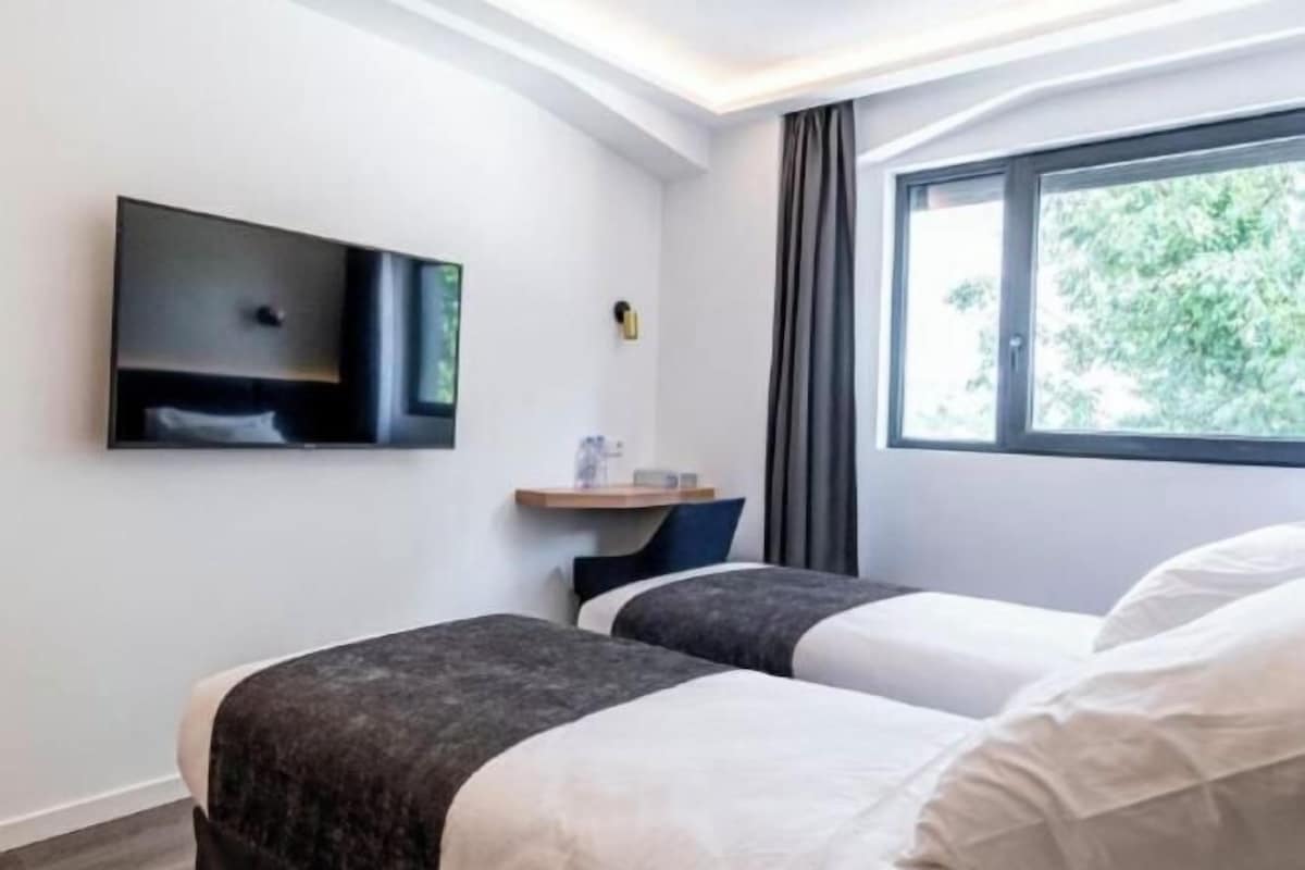 Habitación de hotel con dos camas, un televisor montado en la pared, una ventana con cortinas y una mesita.