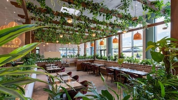 Interior de restaurante com vigas de madeira, plantas penduradas e amplas janelas. Mesas e cadeiras dispostas para refeições.