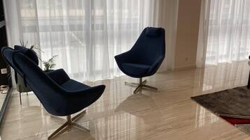 Ein modernes Wohnzimmer mit zwei dunkelblauen Sesseln, einem Glastisch und einem großen Fenster mit transparenten Vorhängen.