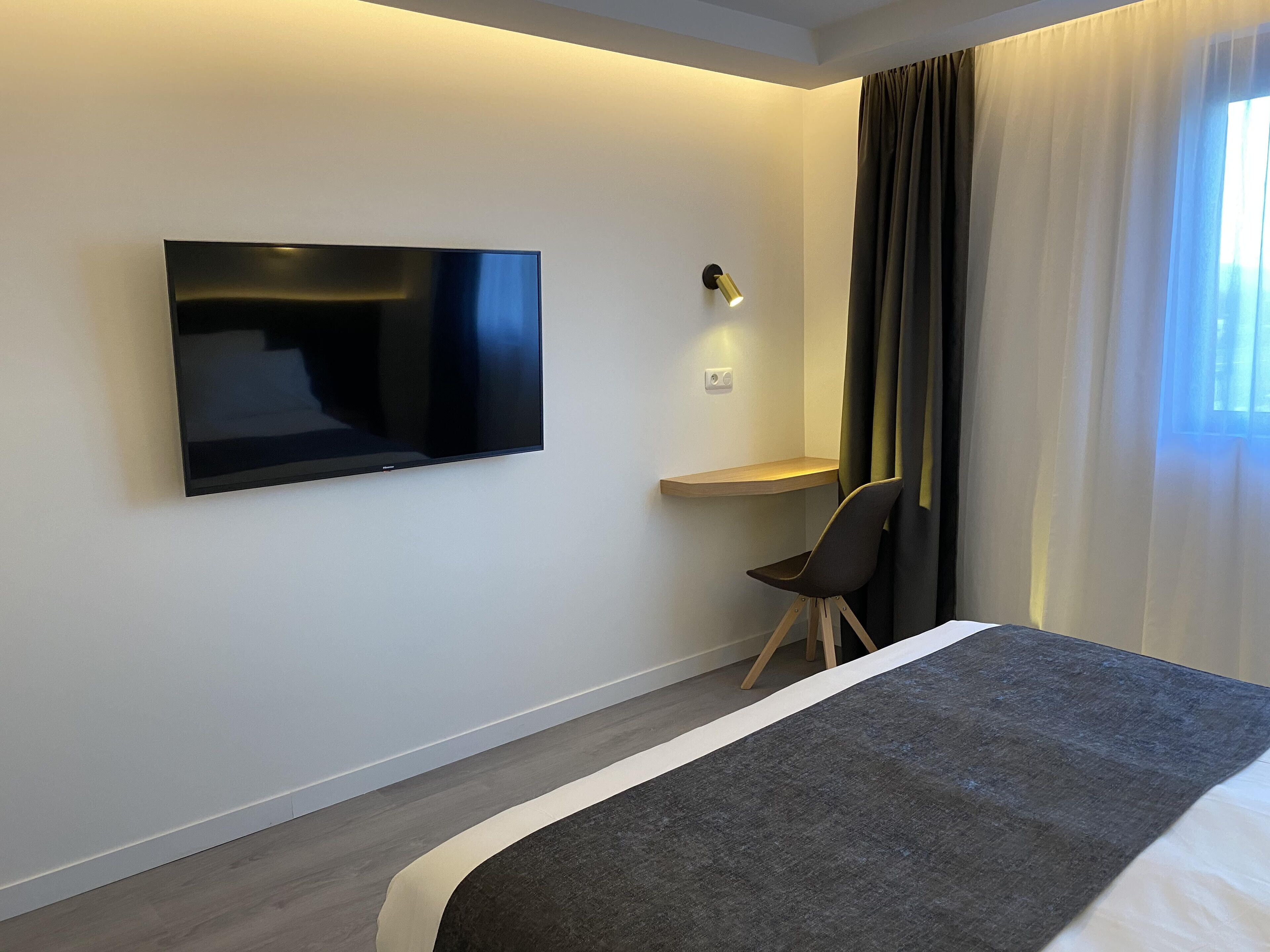 A modern hotel room with a flat-screen TV mounted on the wall, a small desk with a chair, and a large bed with a dark headboard.