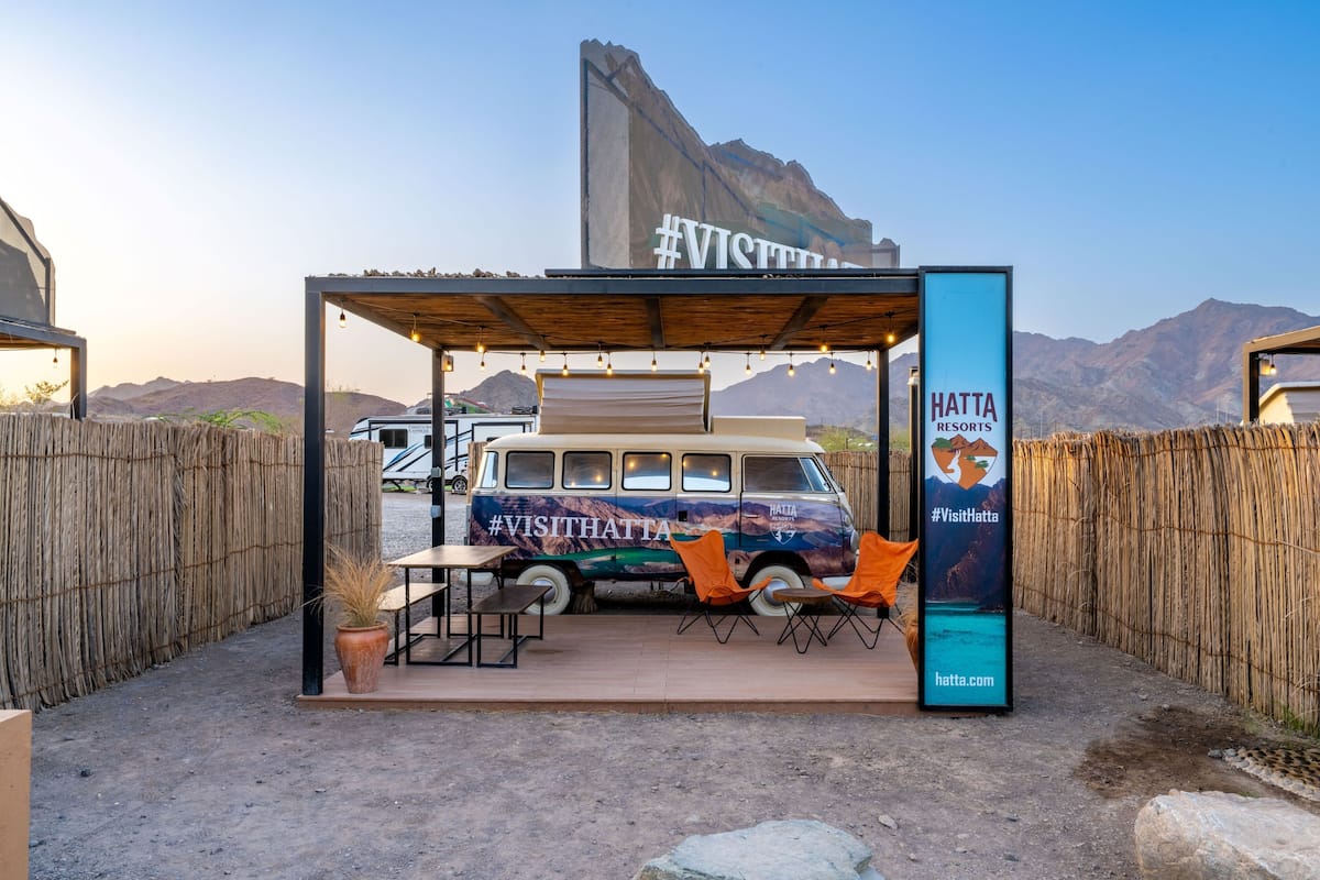 A rustic outdoor seating area with a van and tables, surrounded by a wooden fence and mountains in the background.