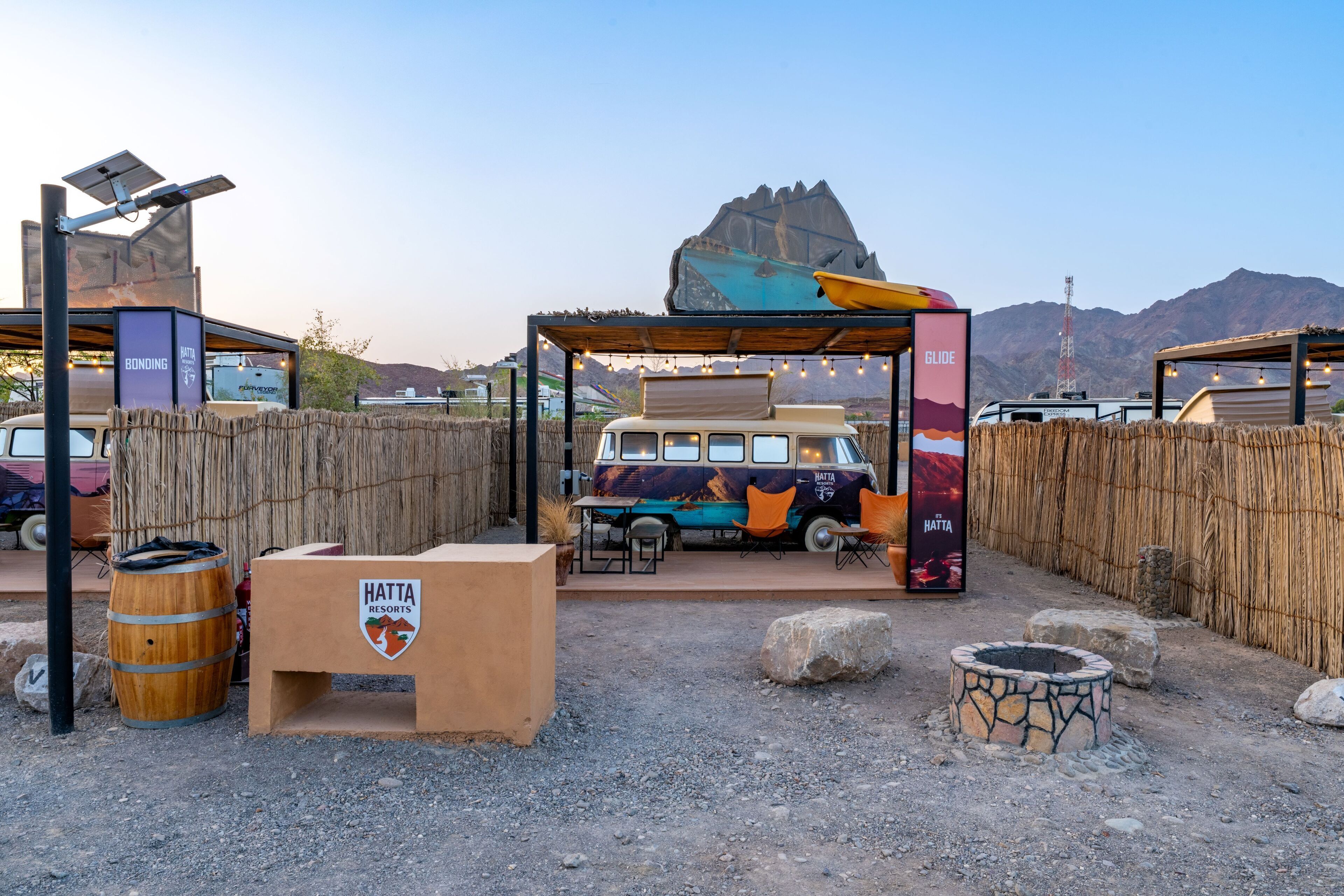 A rustic outdoor seating area with a van, wooden barrels, and a stone fire pit.