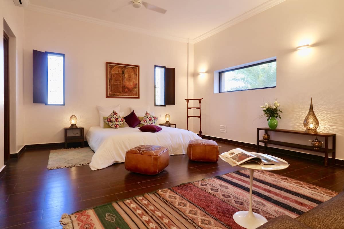 A modern bedroom with a large bed, two ottomans, a standing lamp, a small table with a magazine, and a decorative vase.