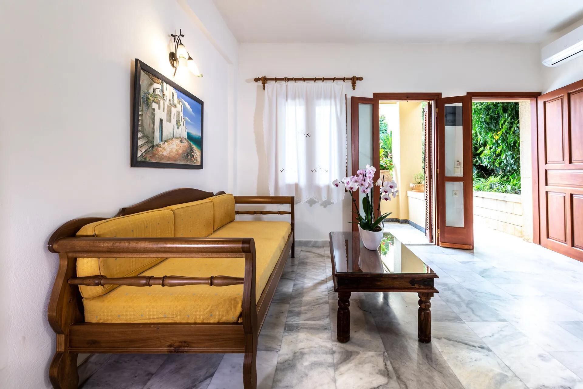A room with a wooden sofa, a small table with a potted orchid, a painting on the wall, and a view of a garden through an open door.