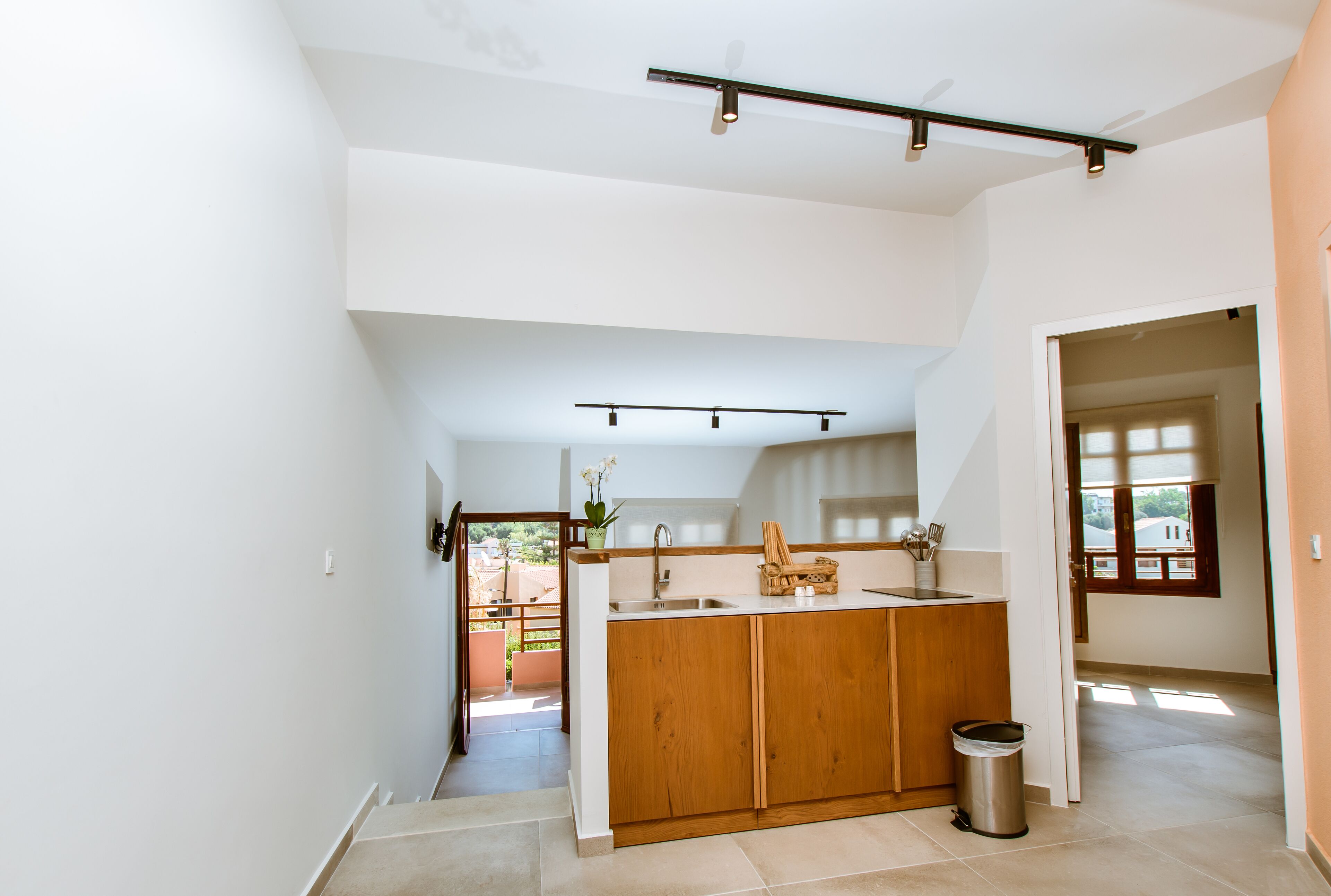 A modern kitchen with wooden cabinets, a countertop, and a sink. There is a trash can and a window providing natural light.