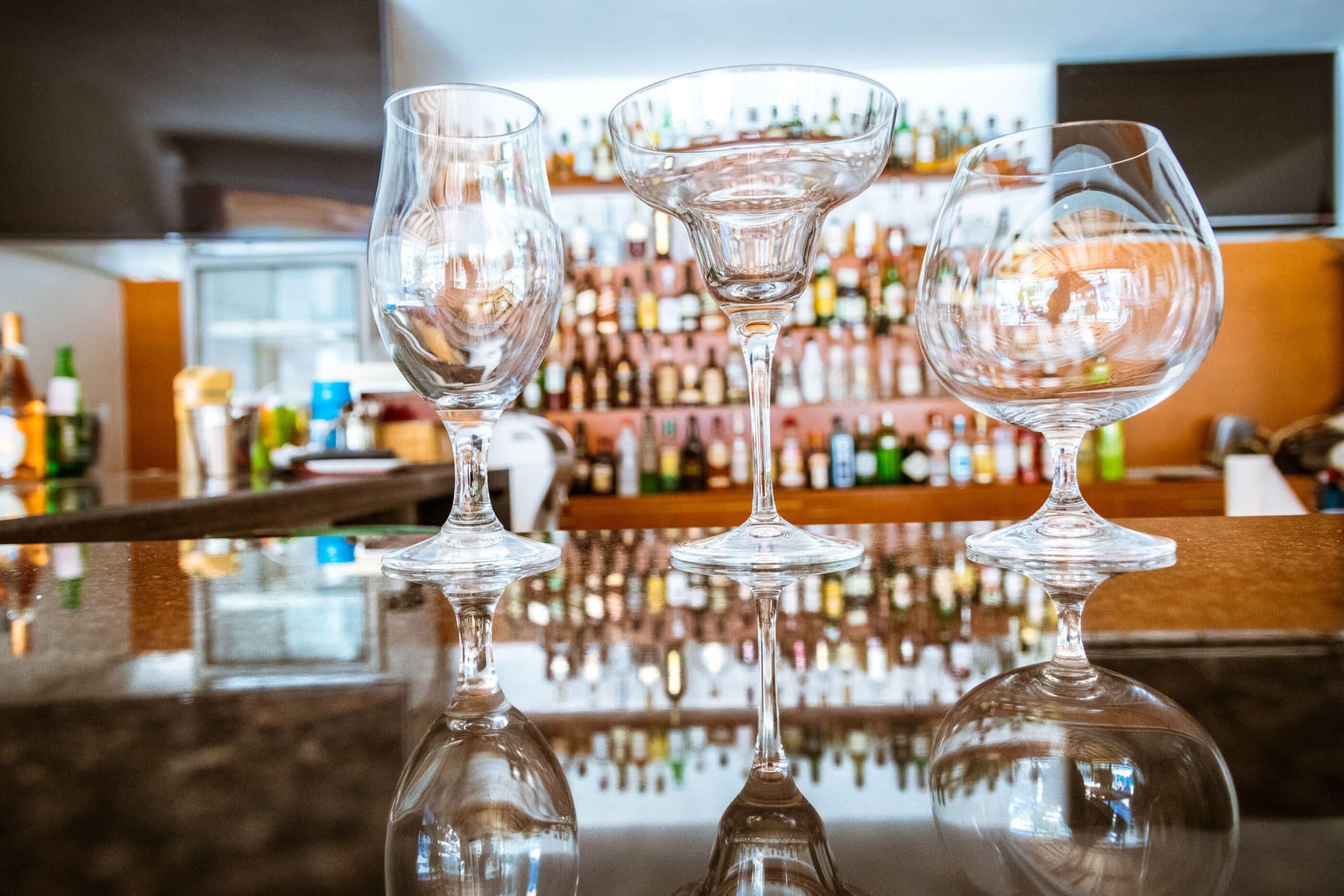 Three empty wine glasses on a bar counter with a reflection of the bar and bottles in the background.