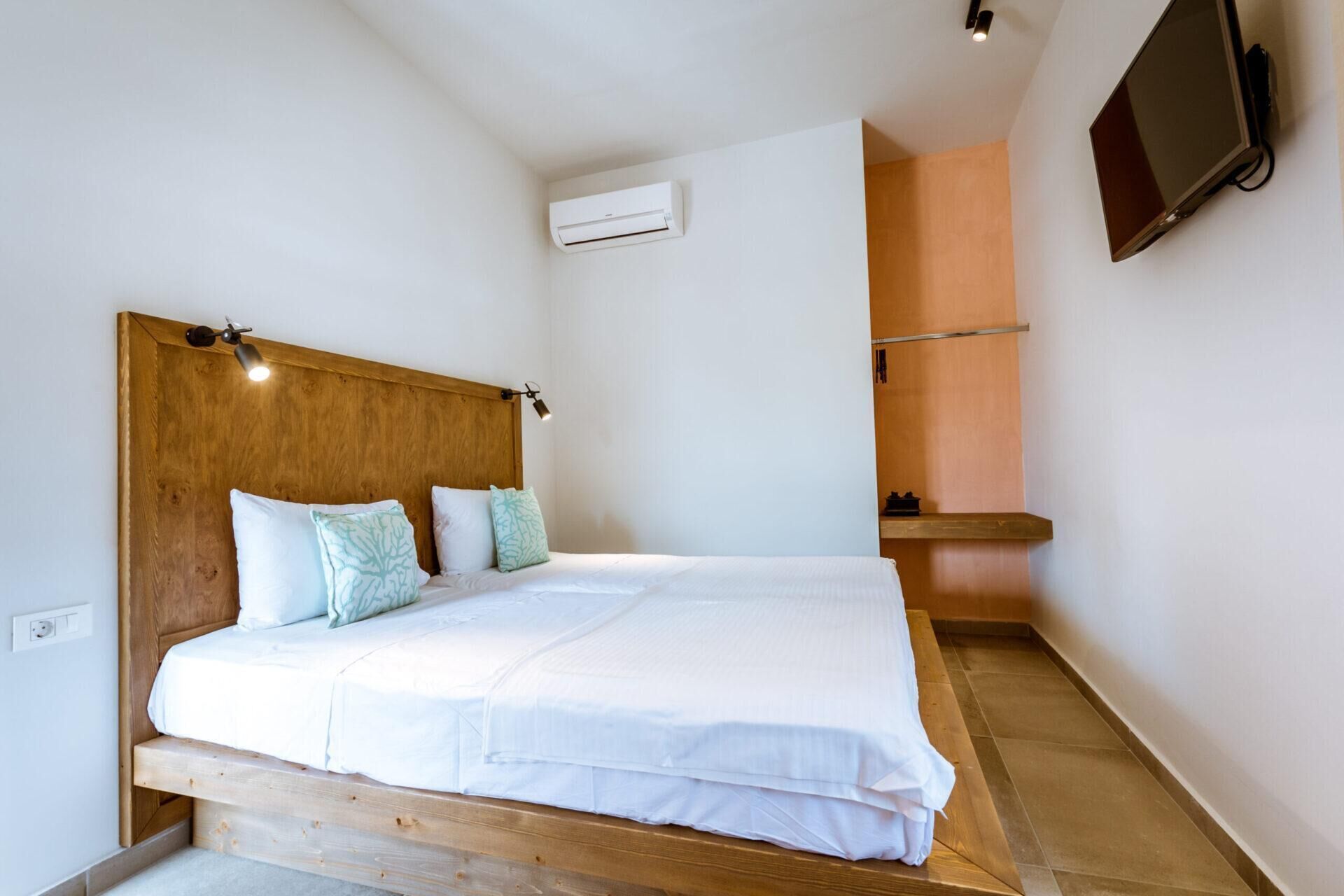 A bedroom with a wooden headboard, a bed with white bedding, an air conditioning unit, a flat-screen TV, and a small wooden shelf.