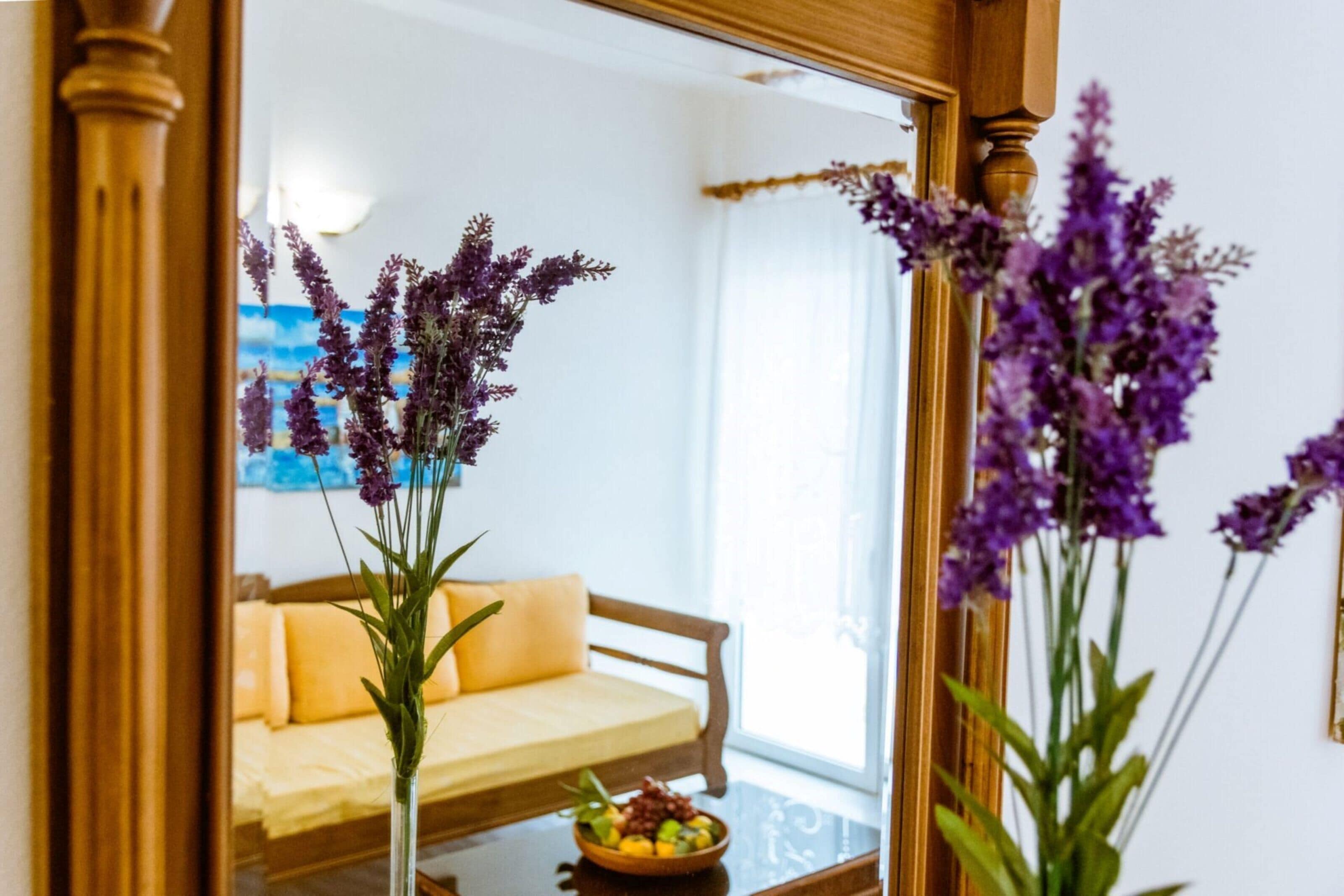 A room with a bed, a mirror, and a vase with purple flowers.