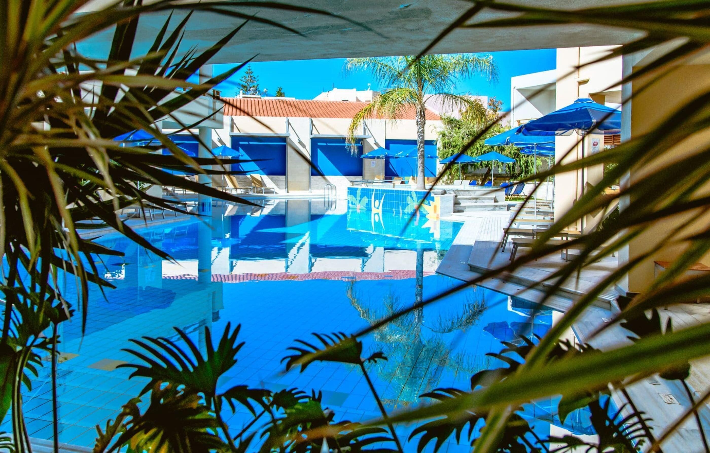 A clear view of a swimming pool with lounge chairs and umbrellas, surrounded by palm trees and buildings.