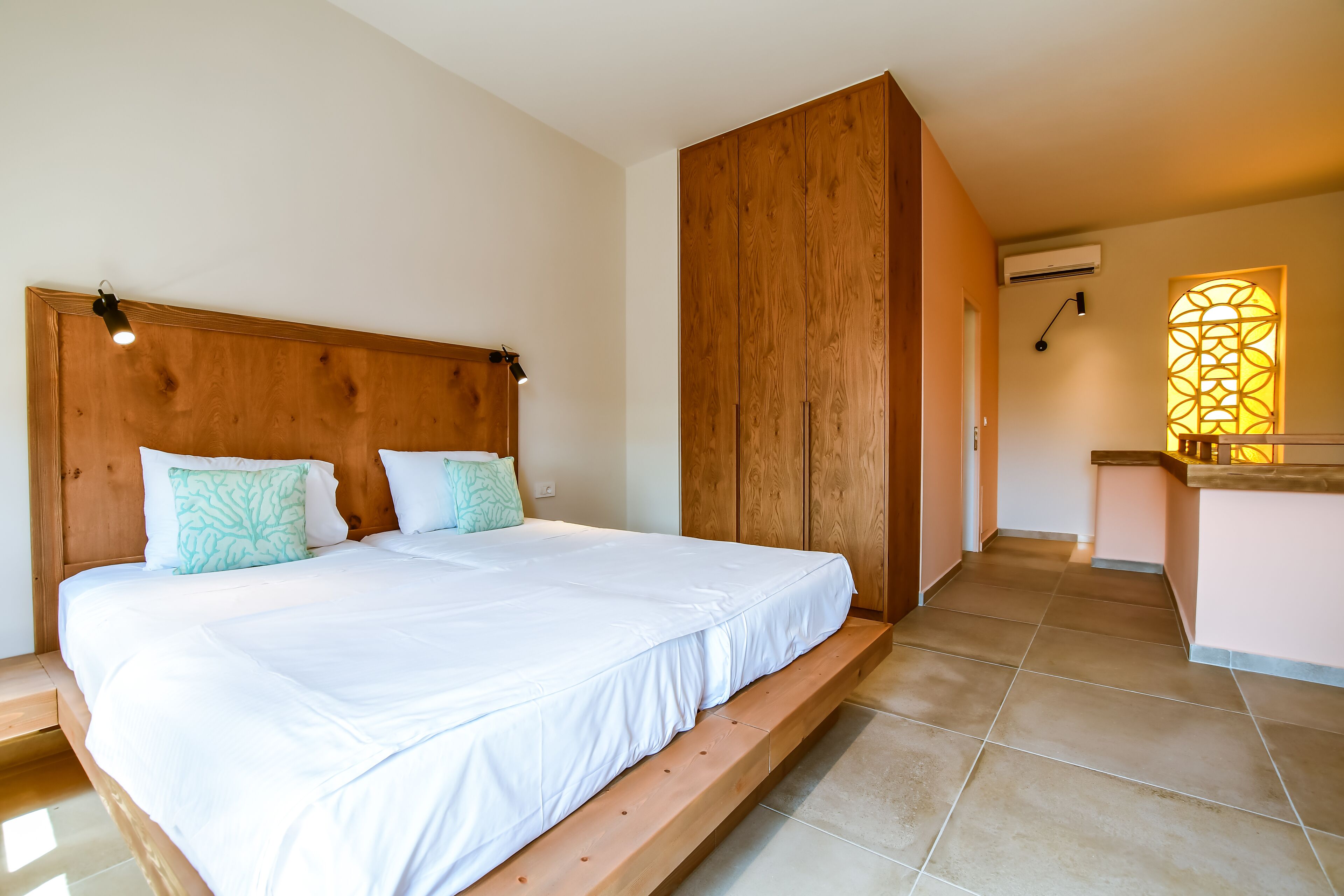 A bedroom with a wooden headboard, a large bed with white bedding, and a small bathroom with a wooden counter and a window.