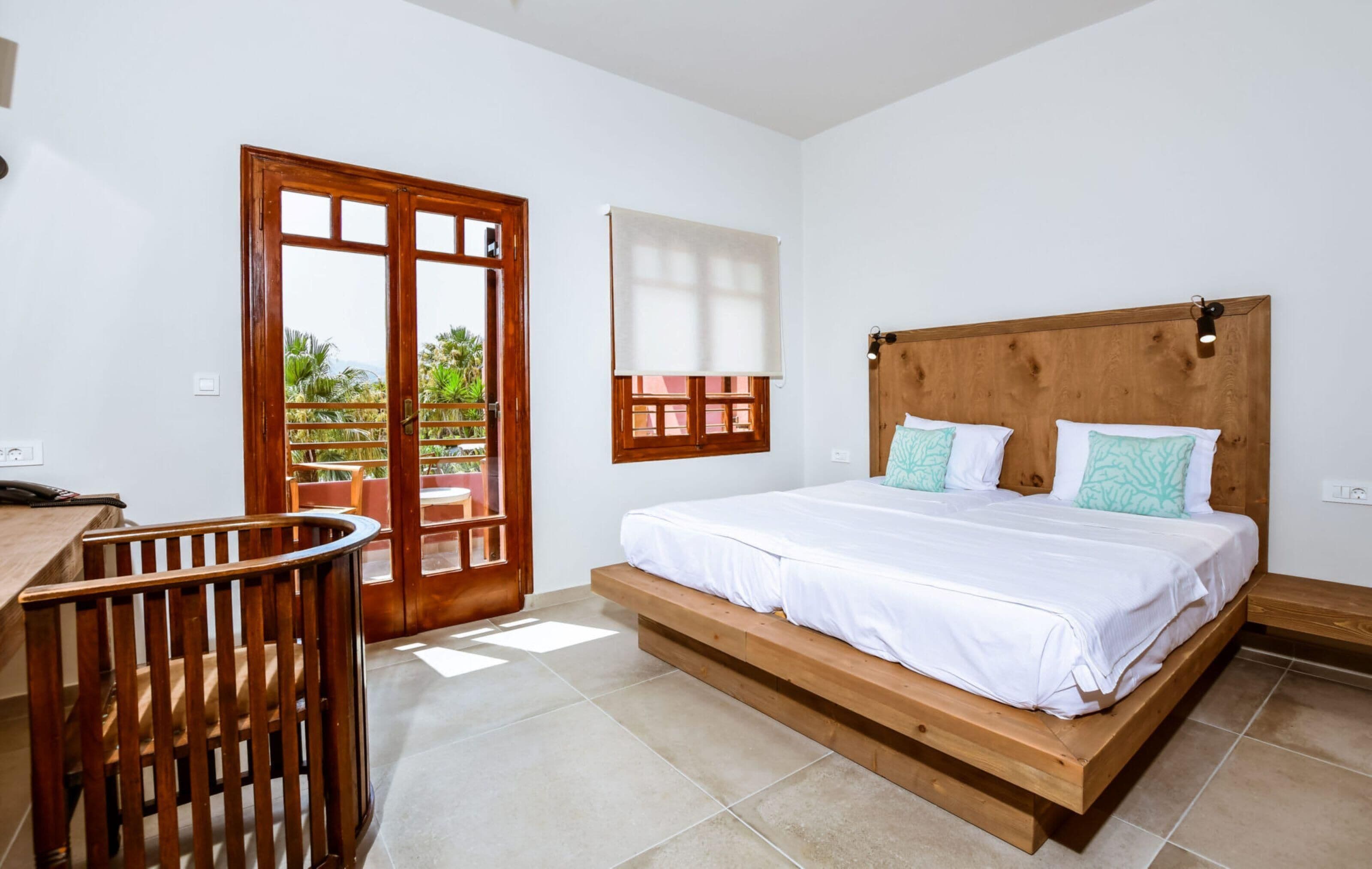 A bedroom with a wooden bed, a desk, and a chair. There is a window with a view of greenery outside.