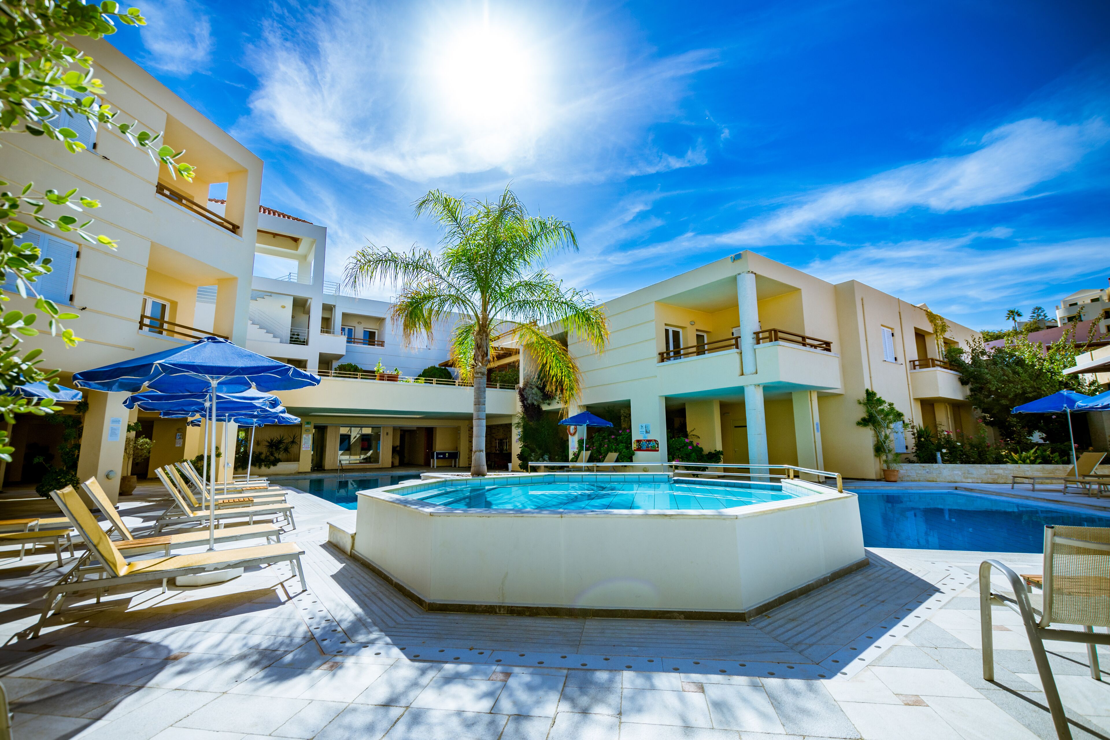Zona de piscina con tumbonas y sombrillas, rodeada de edificios con balcones.
