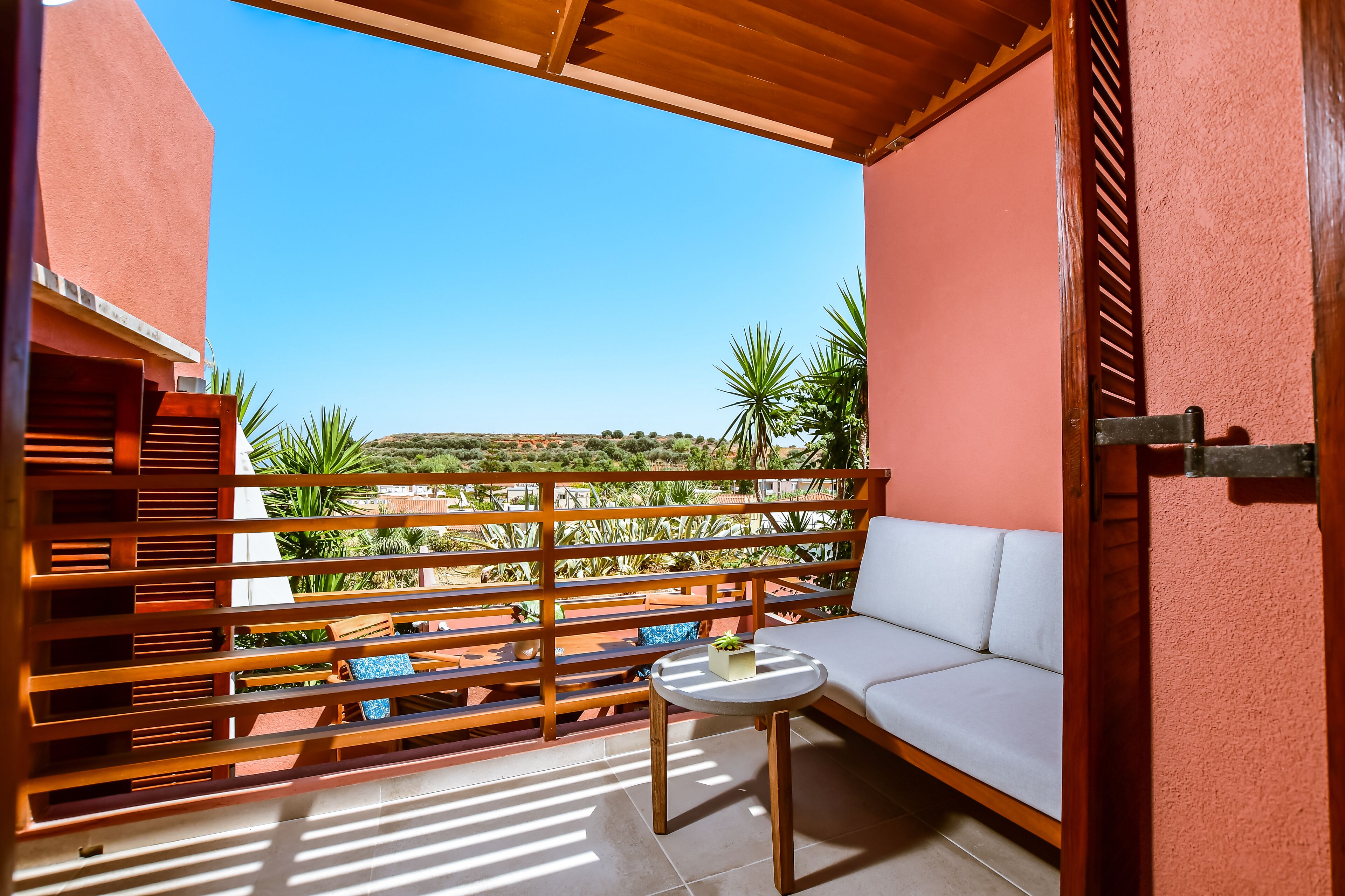 Un balcón con sofá blanco, una mesita pequeña, barandillas de madera y vistas a zonas verdes y colinas.