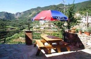 A wooden picnic table with a red and blue umbrella labeled "VATI" in a mountainous area with stone walls and buildings in the background. - Sandra Villa Rooms (Vernazza)