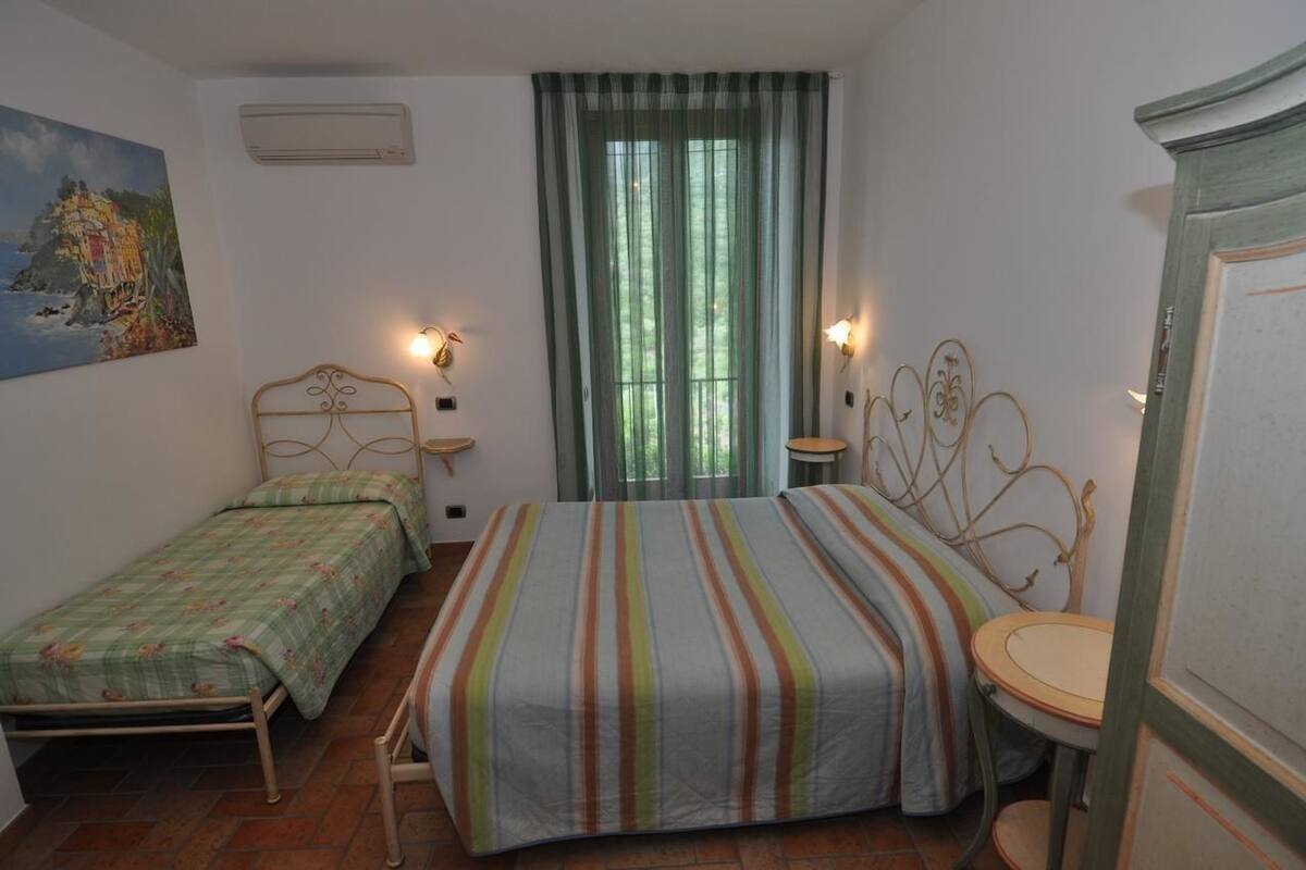 A bedroom with two beds, a window with curtains, a painting on the wall, and a small table with a chair.
