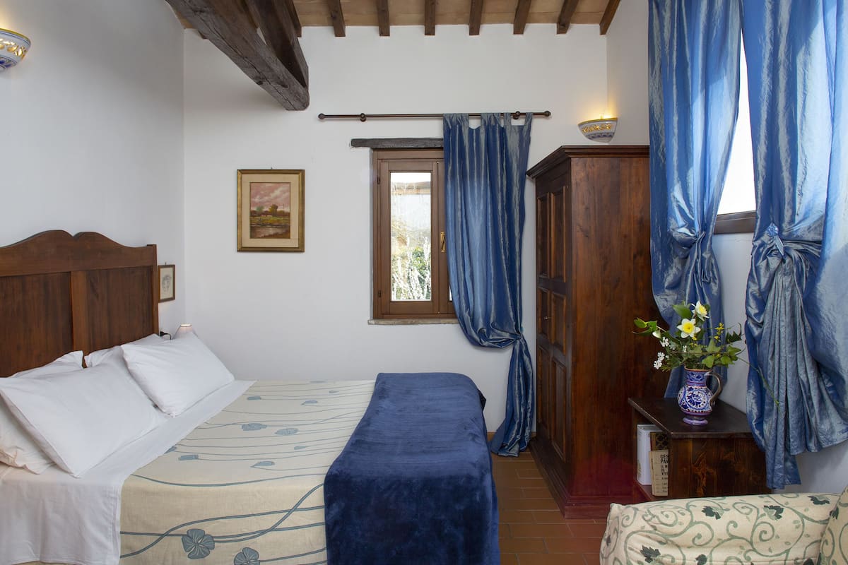 A bedroom with a bed, a bedside table with a vase, a window with curtains, and a wooden wardrobe.