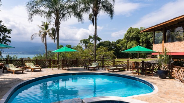 Ein Swimmingpool mit Liegestühlen und Sonnenschirmen, umgeben von Palmen und Blick aufs Meer.