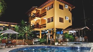 Ein mehrstöckiges Hotel mit Poolbereich, Sitzgelegenheiten im Freien und tropischer Bepflanzung.