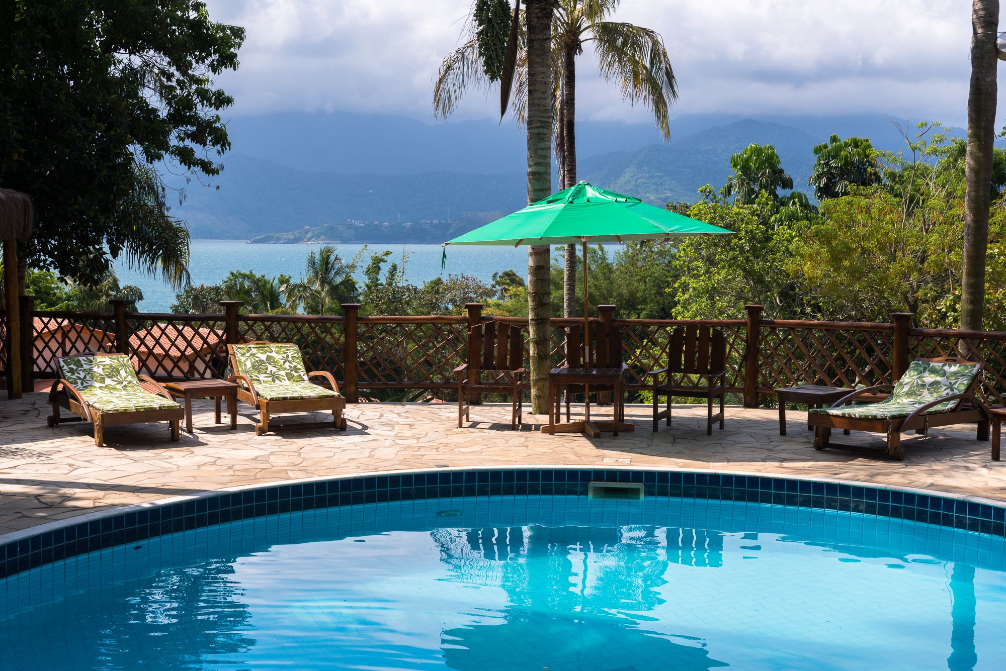 Zona de piscina con sillones de descanso y una sombrilla, rodeada por una valla de madera y palmeras, con vistas a un cuerpo de agua y montañas.