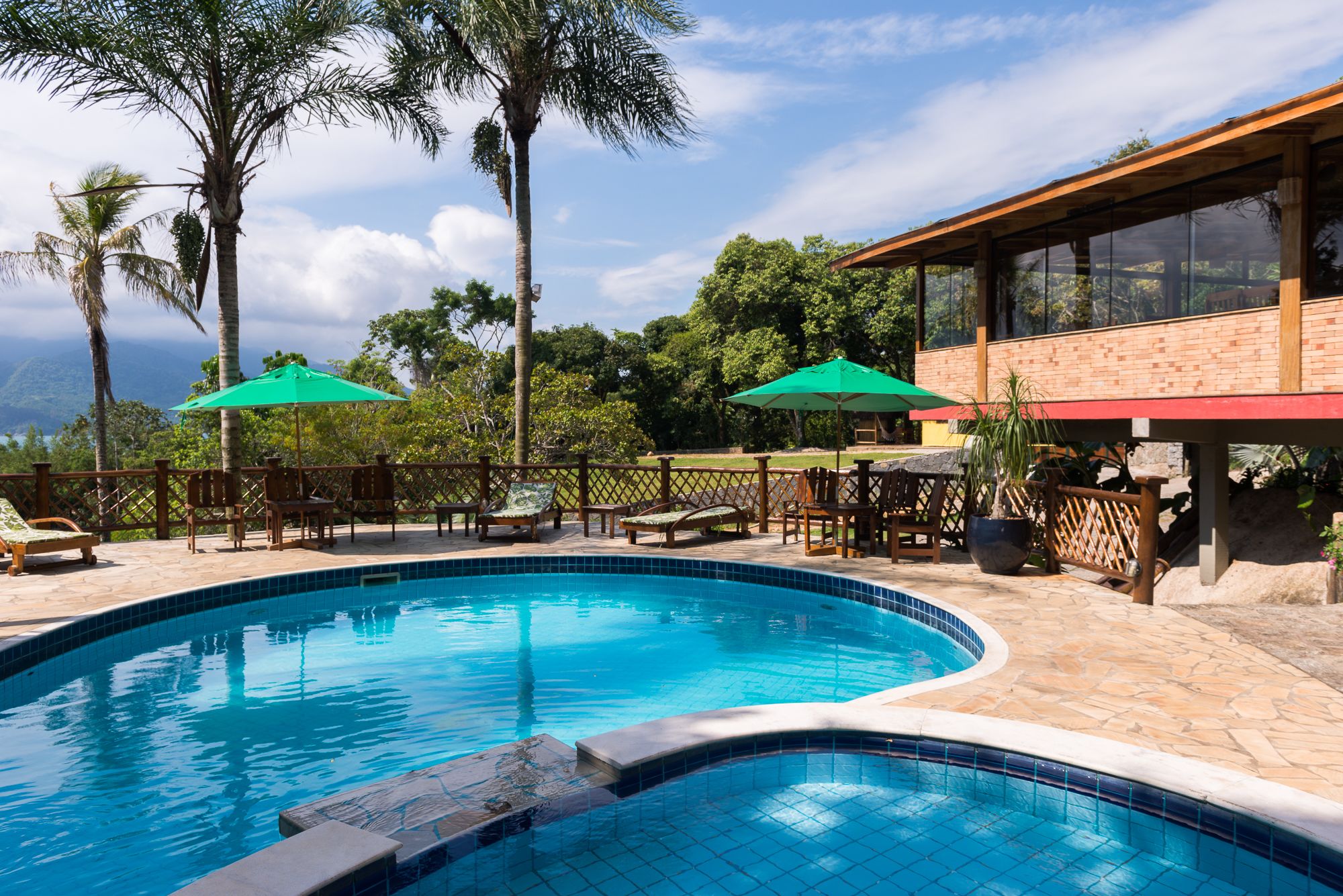 A swimming pool with lounge chairs and umbrellas, surrounded by palm trees and a brick building.