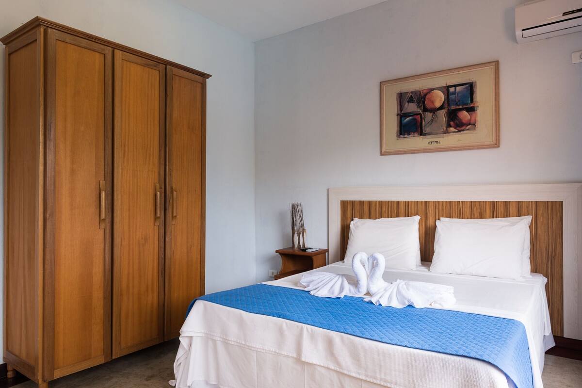 Une chambre d’hôtel avec une grande armoire en bois, un lit avec une literie blanche et bleue, et un tableau accroché au mur.