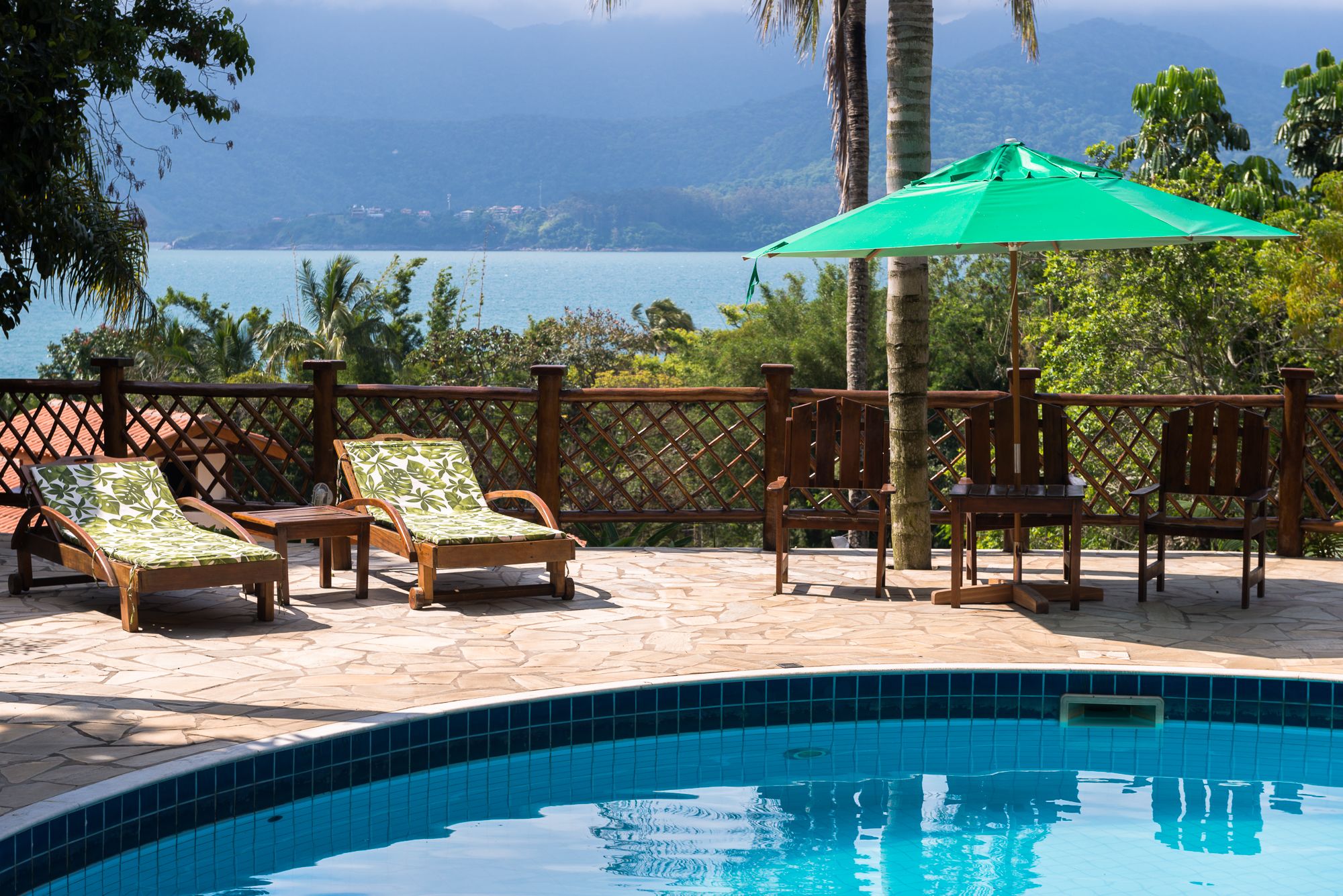 Zona de piscina con sillas de descanso y una sombrilla verde, rodeada por una valla de madera y palmeras, con vista a una masa de agua.